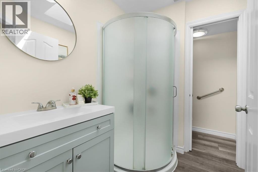 Vanity with integrated white basin and chrome faucet, curved glass shower enclosure, wood-finish flooring, and a wall-mounted oval mirror - 150 Bankside Drive, Kitchener, ON