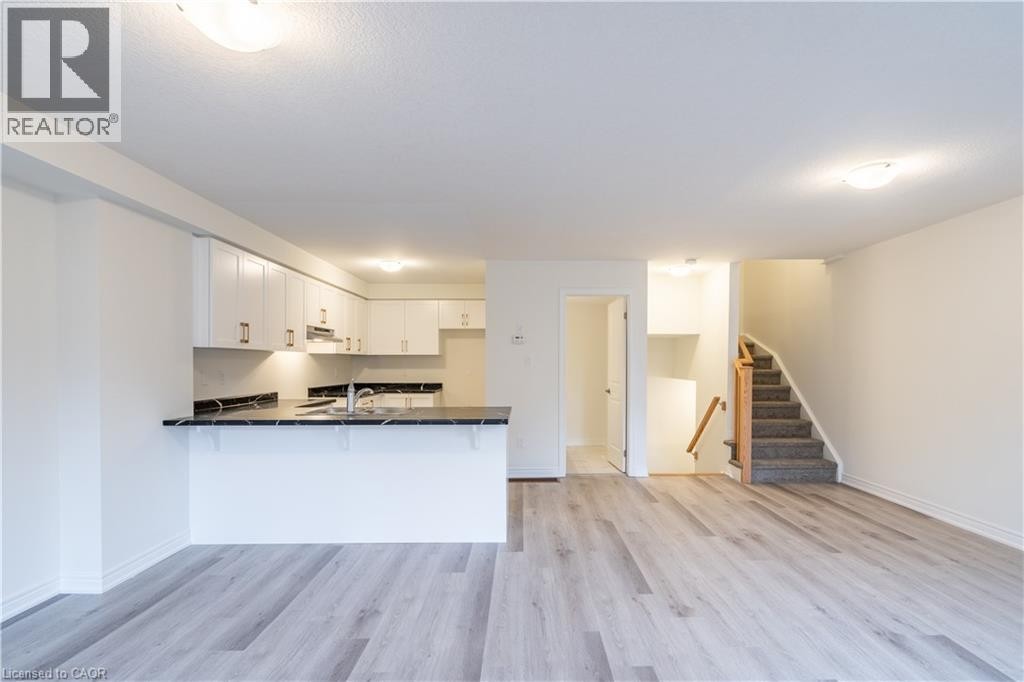 Light wood-finish flooring throughout, white upper and lower cabinetry, dark countertops, and a carpeted staircase with wood handrail - 23 Dryden Lane, Hamilton, ON