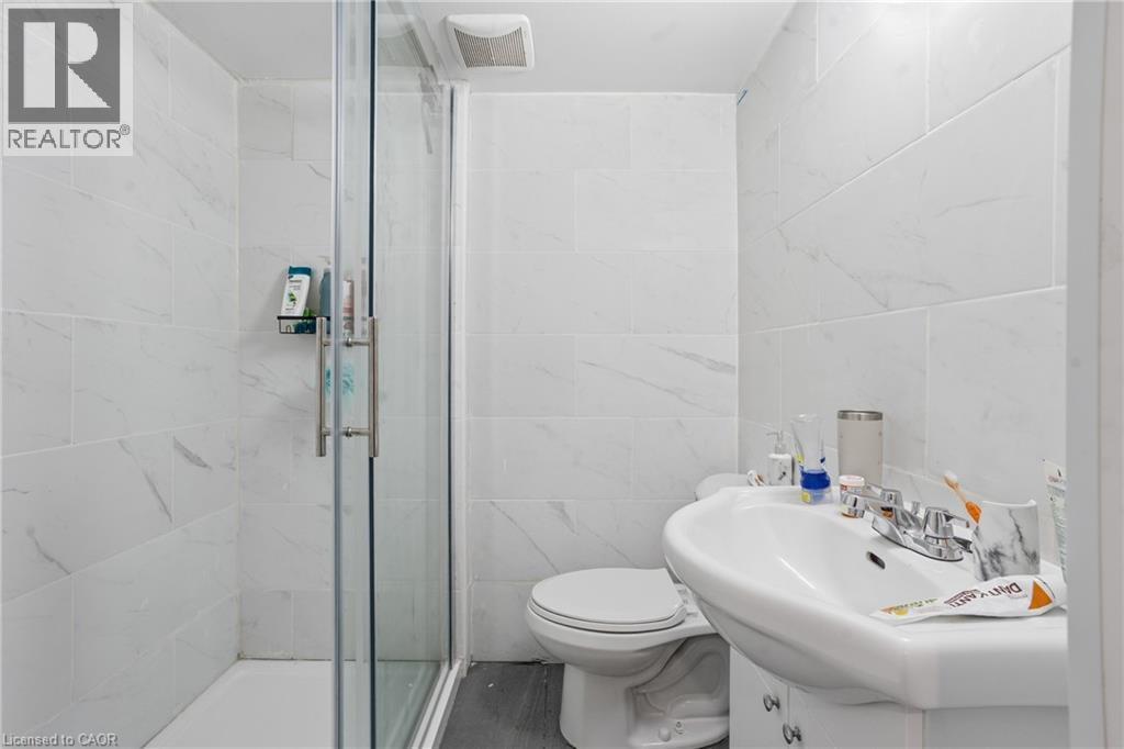 Bathroom featuring a glass-enclosed shower, white ceramic wall tiles with subtle veining, grey wood-finish flooring, a pedestal sink with chrome fixtures, and an exhaust fan - 139 Dalewood Avenue, Hamilton, ON