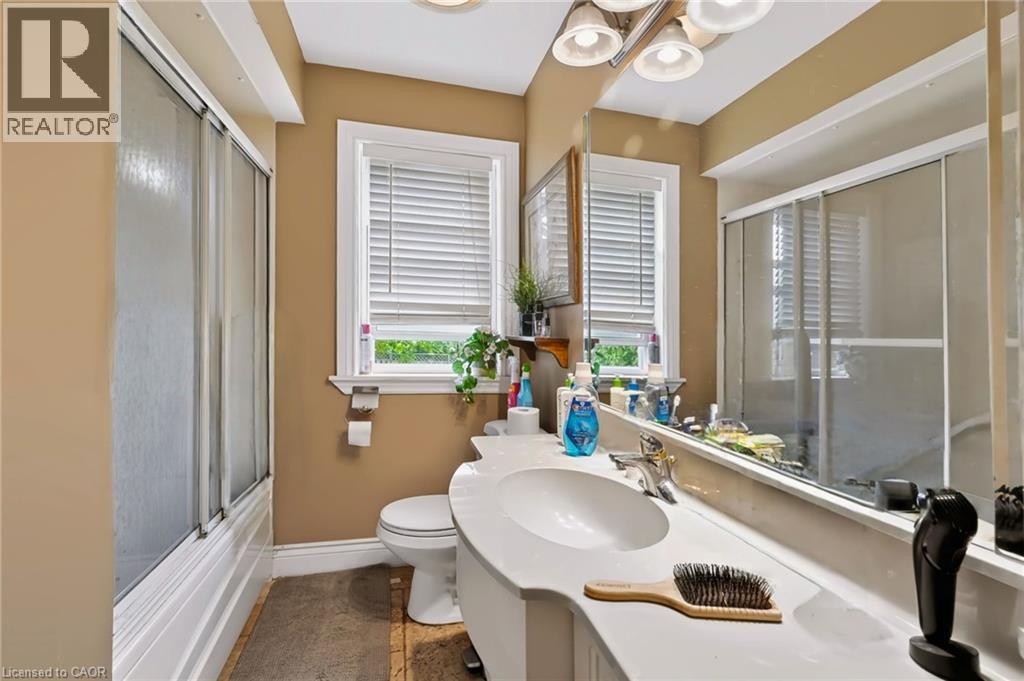Bathroom featuring a single vanity with an integrated sink, broad wall-mounted mirror, a window with horizontal blinds, and a shower-tub combination with sliding glass doors - 1046 Shepherds Drive, Burlington, ON