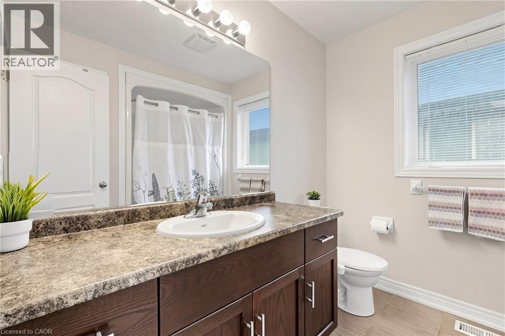 Bathroom featuring a wood-finish vanity with a stone-look countertop, an integrated sink, a full-width mirror, and a window with blinds - 173 Raymond Road, Ancaster, ON