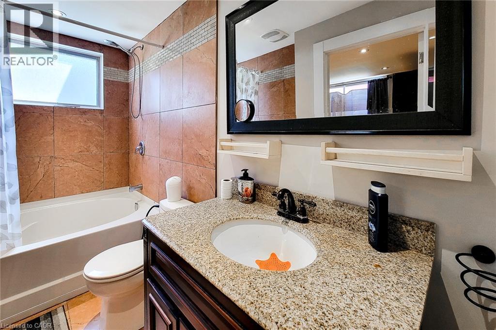 Integrated bathtub and shower with brown tile surround, a frosted glass window, a vanity with a granite-style countertop, and a dark wood-finish cabinet - 1 Vera Court, Hamilton, ON