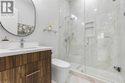 Modern bathroom featuring a wood-finish vanity with a white countertop, a frameless oval mirror, a glass-enclosed shower with marble-look wall tiles, and an integrated shower niche - 