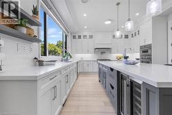 Spacious kitchen featuring an expansive central island with integrated beverage refrigerator, light wood-finish flooring, white perimeter cabinetry, stainless steel appliances, and a large window abov - 