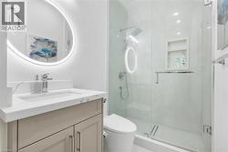 Modern bathroom featuring a backlit circular mirror, a vanity with a light-toned countertop, a frameless glass shower enclosure, a built-in shower niche, and a contemporary shower fixture - 