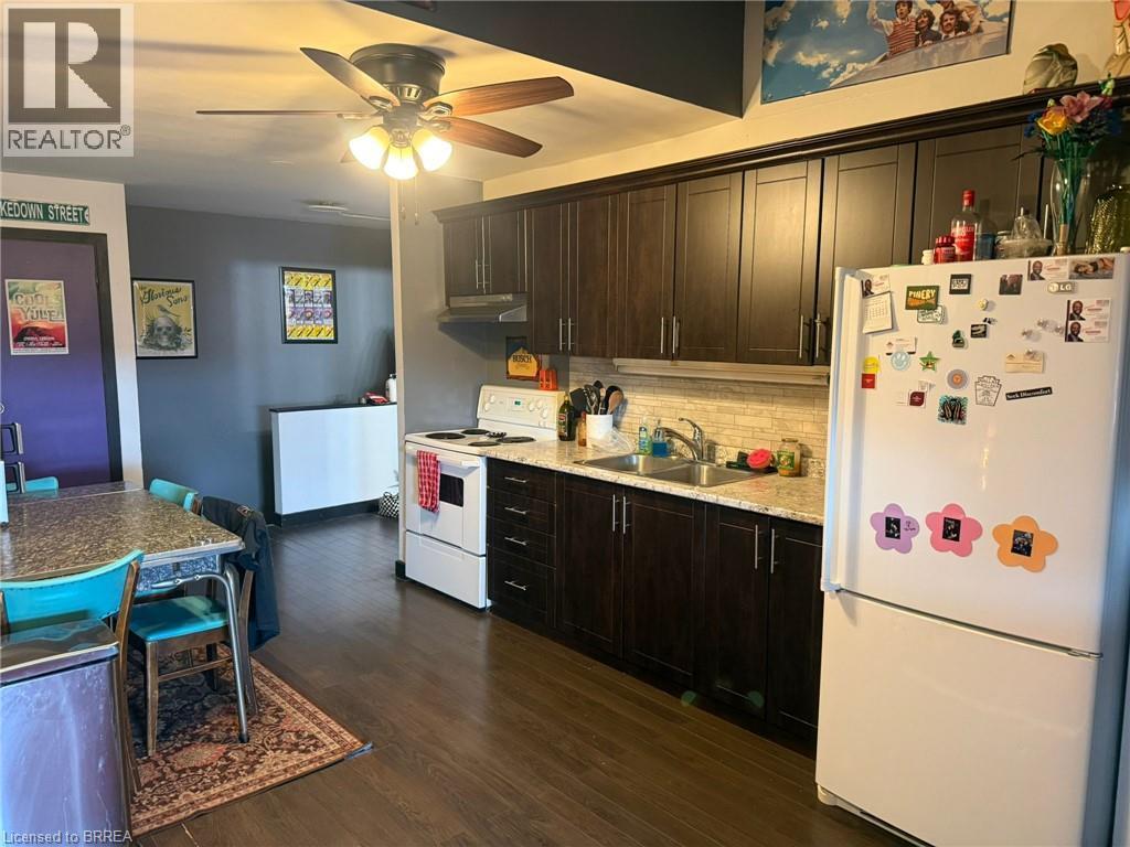 Contemporary kitchen featuring dark cabinetry with brushed metal hardware, white appliances including a refrigerator and electric range, wood-finish flooring, and a ceiling fan with integrated lightin - 12 Bridgeport Road E Unit# 18, Waterloo, ON