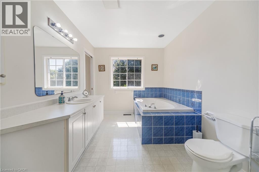 Dual window placement, built-in bathtub with blue tile surround, vanity with integrated sink, rectangular mirror with overhead lighting, and white toilet - 82 Milner Downs Crescent, Ottawa, ON