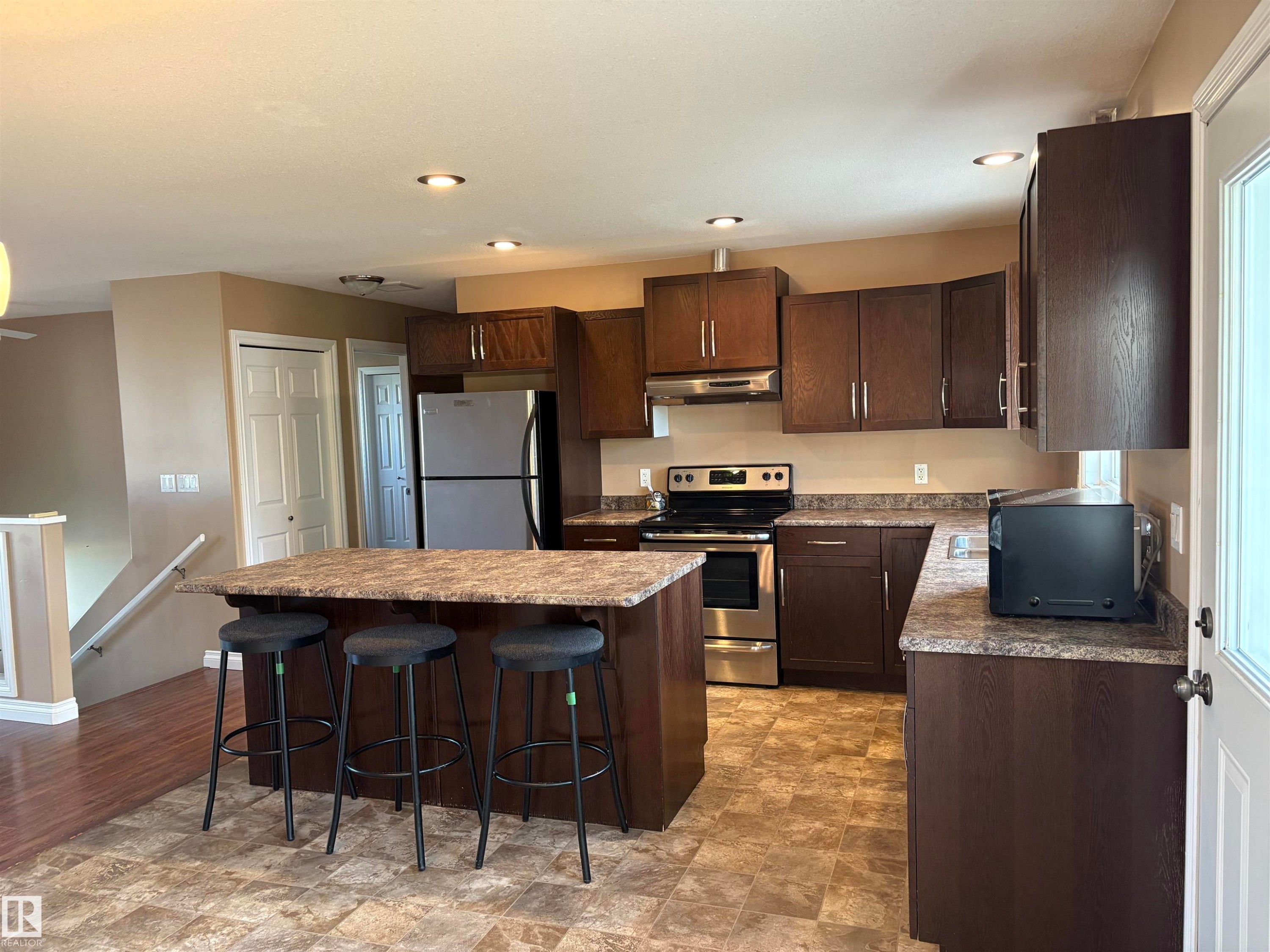 Kitchen with dark brown cabinets, a kitchen bar, appliances with stainless steel finishes, a kitchen island, and under cabinet range hood - 5302 Railway Avenue, Elk Point, AB - Indoor Photo Showing Kitchen