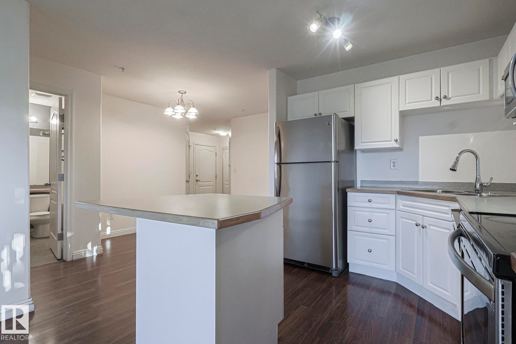 Kitchen featuring freestanding refrigerator, white cabinets, a center island, dark wood-style floors, and pendant lighting - 120 7511 171 Street Nw, Edmonton, AB - Indoor Photo Showing Kitchen