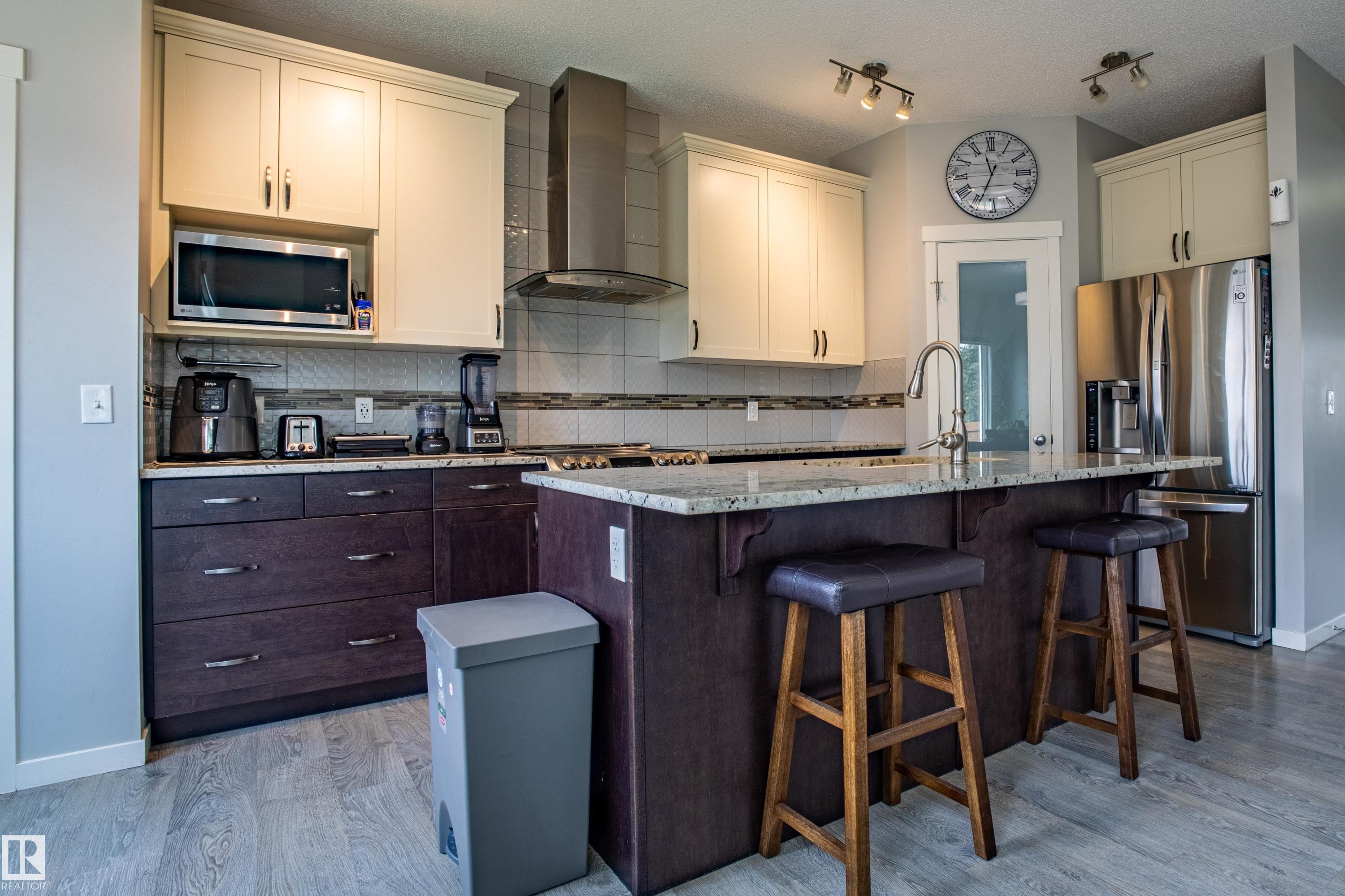 Kitchen with wall chimney range hood, stainless steel appliances, backsplash, light stone countertops, and a textured ceiling - 2407 Casey Link, Edmonton, AB - Indoor Photo Showing Kitchen