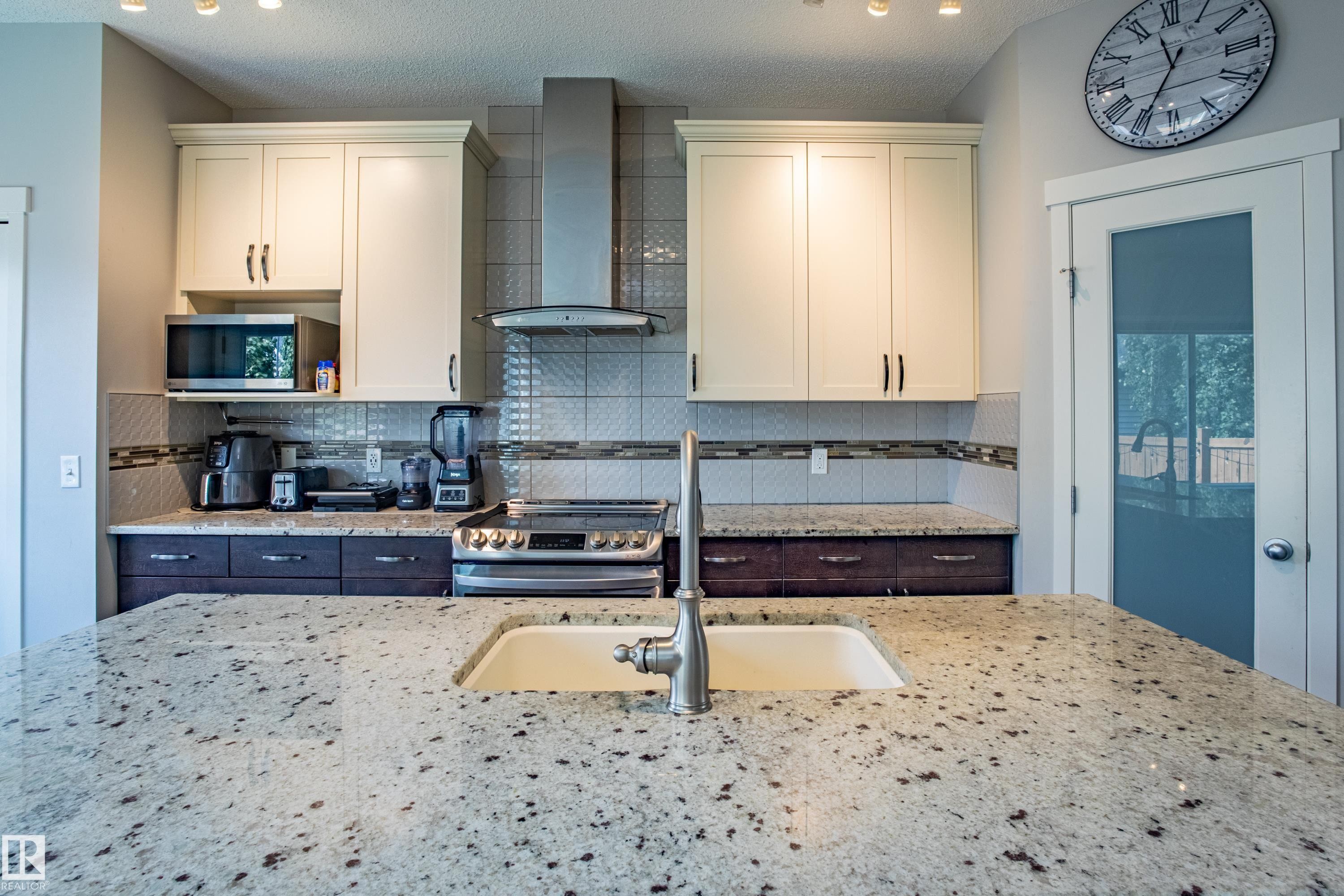 Kitchen featuring wall chimney exhaust hood, appliances with stainless steel finishes, light stone countertops, backsplash, and a textured ceiling - 2407 Casey Link, Edmonton, AB - Indoor Photo Showing Kitchen With Double Sink