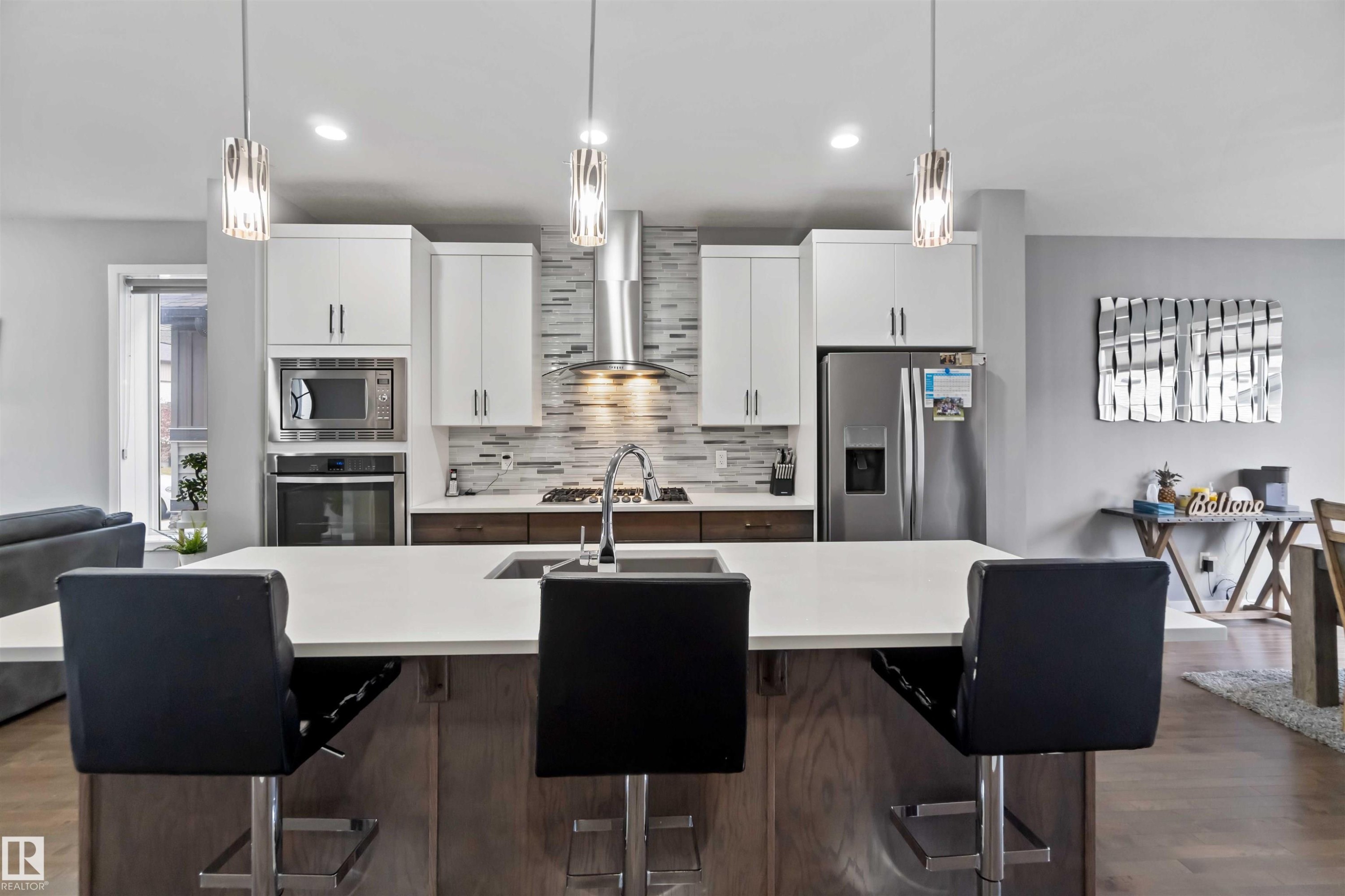 Kitchen featuring backsplash, appliances with stainless steel finishes, a breakfast bar area, white cabinetry, and wall chimney range hood - 17816 59 Street, Edmonton, AB