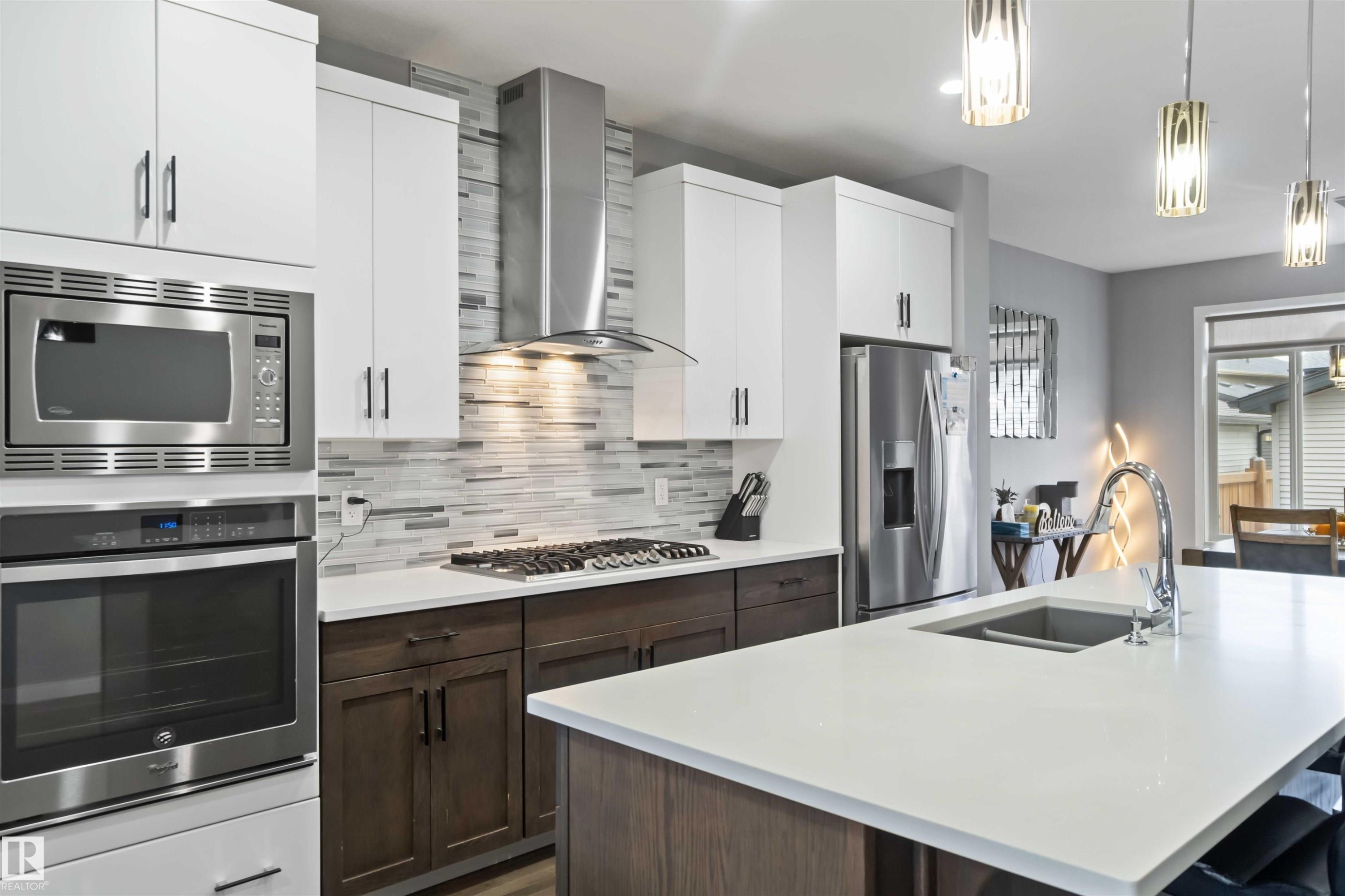 Kitchen featuring decorative backsplash, appliances with stainless steel finishes, pendant lighting, wall chimney range hood, and an island with sink - 17816 59 Street, Edmonton, AB