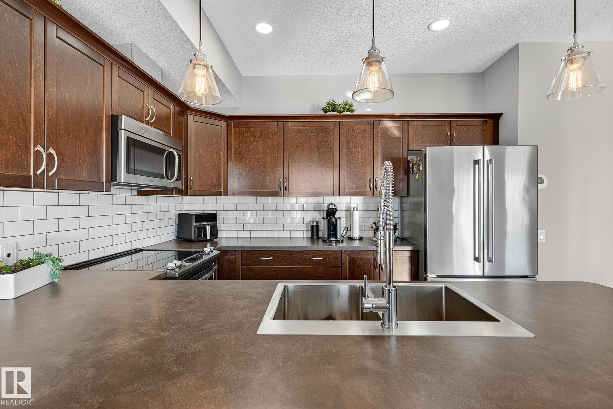 Kitchen featuring dark countertops, appliances with stainless steel finishes, backsplash, hanging light fixtures, and a textured ceiling - 117 7293 South Terwillegar Drive, Edmonton, AB - Indoor Photo Showing Kitchen With Double Sink