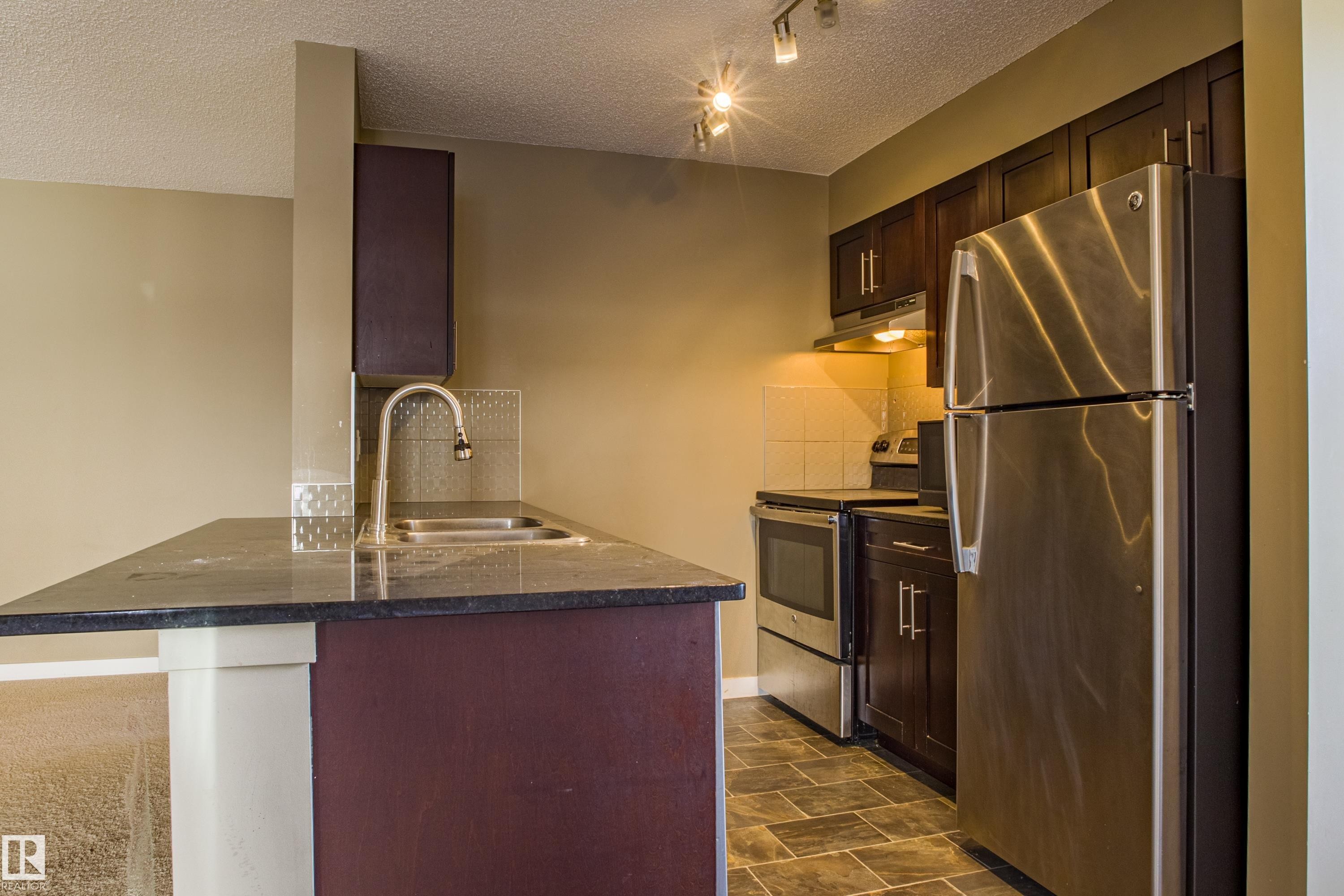 Kitchen with appliances with stainless steel finishes, dark brown cabinets, a textured ceiling, under cabinet range hood, and decorative backsplash - 201 5804 Mullen Place, Edmonton, AB - Indoor Photo Showing Kitchen With Double Sink
