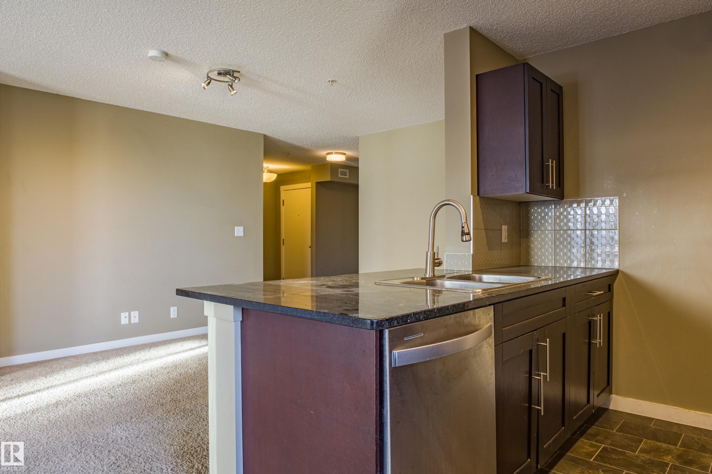 Kitchen featuring dishwasher, dark stone counters, decorative backsplash, a peninsula, and a textured ceiling - 201 5804 Mullen Place, Edmonton, AB - Indoor Photo Showing Kitchen