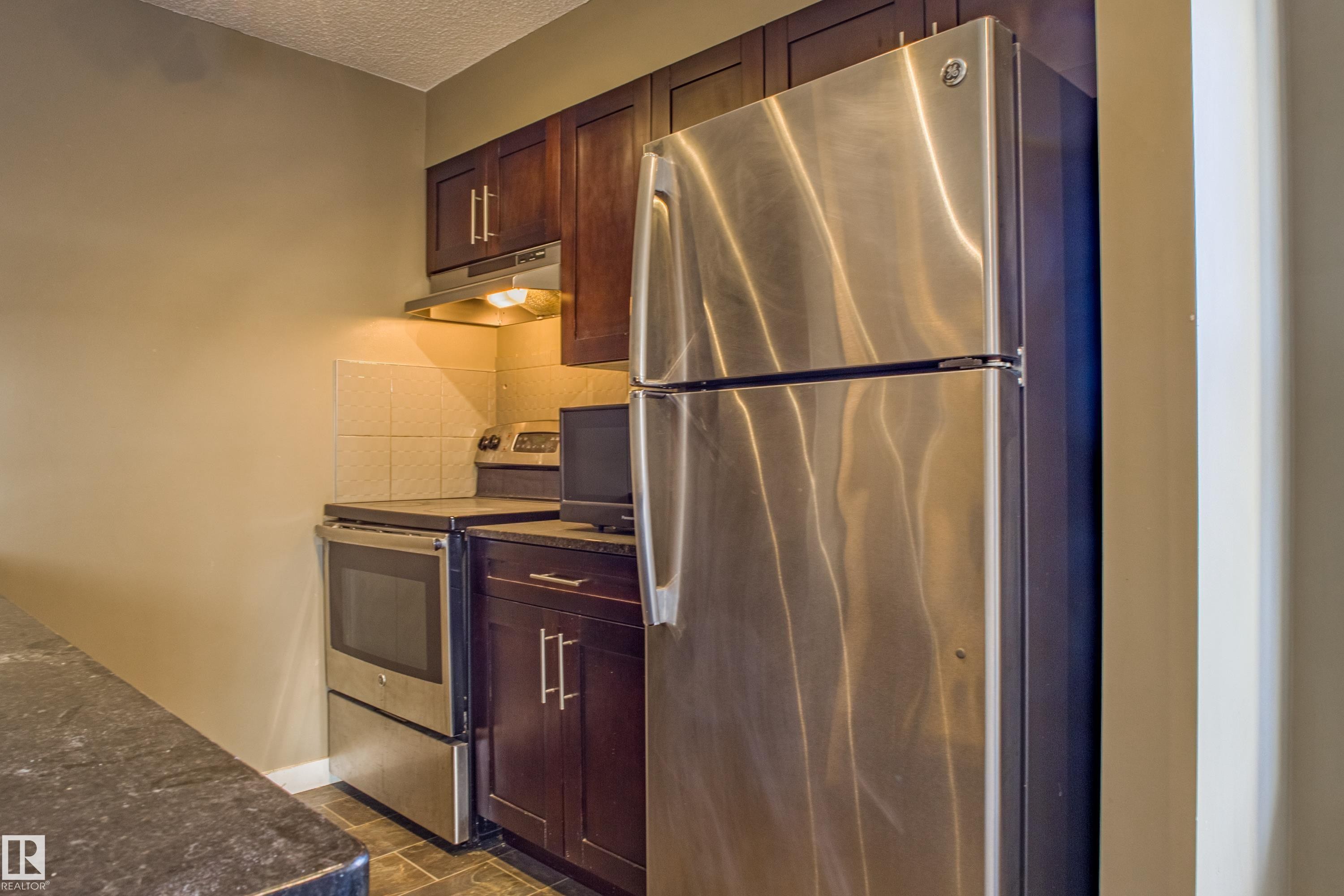 Kitchen featuring stainless steel appliances, under cabinet range hood, a textured ceiling, dark brown cabinetry, and dark stone countertops - 201 5804 Mullen Place, Edmonton, AB - Indoor Photo Showing Kitchen
