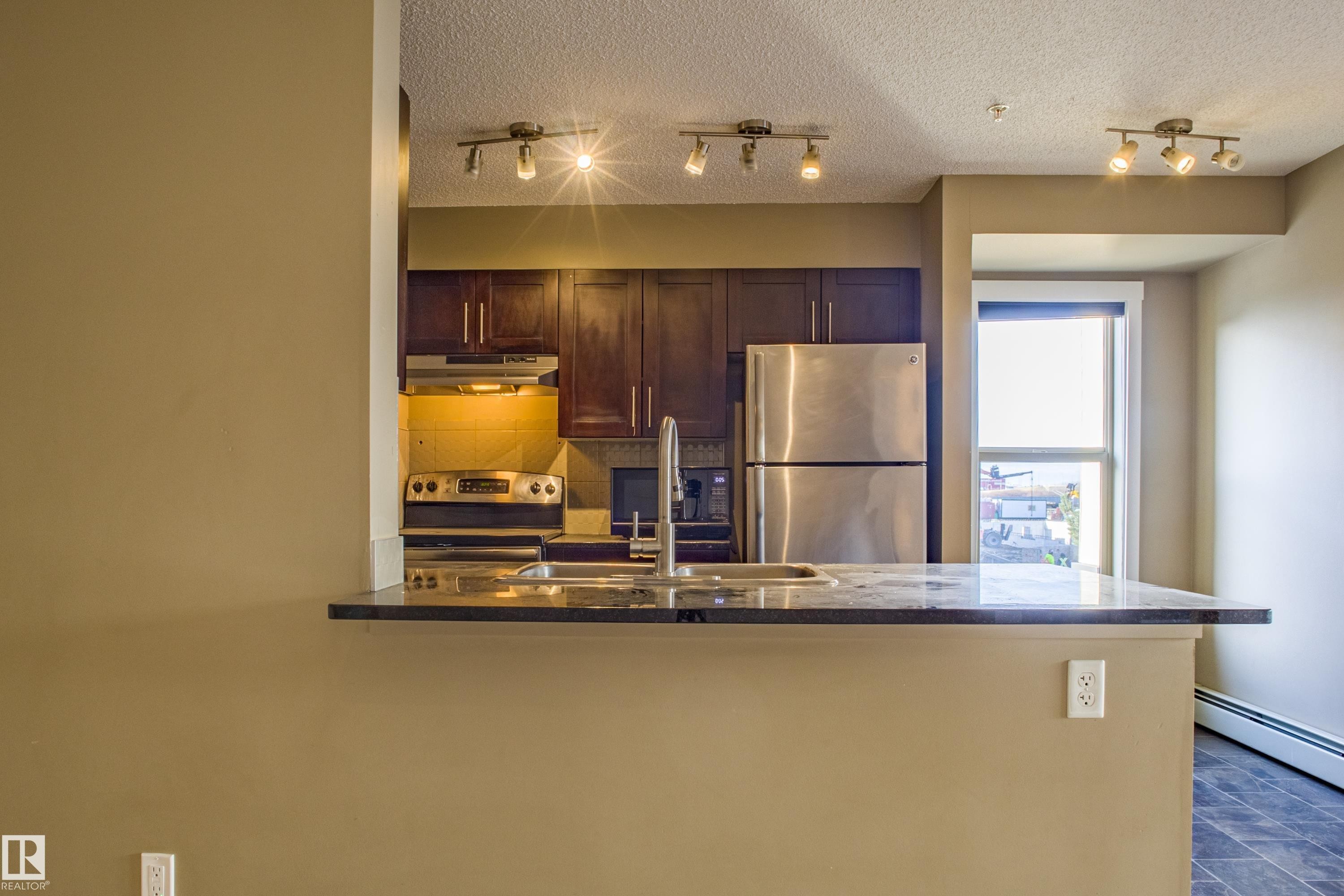 Kitchen featuring tasteful backsplash, stainless steel appliances, a peninsula, under cabinet range hood, and dark stone countertops - 201 5804 Mullen Place, Edmonton, AB - Indoor Photo Showing Kitchen With Double Sink