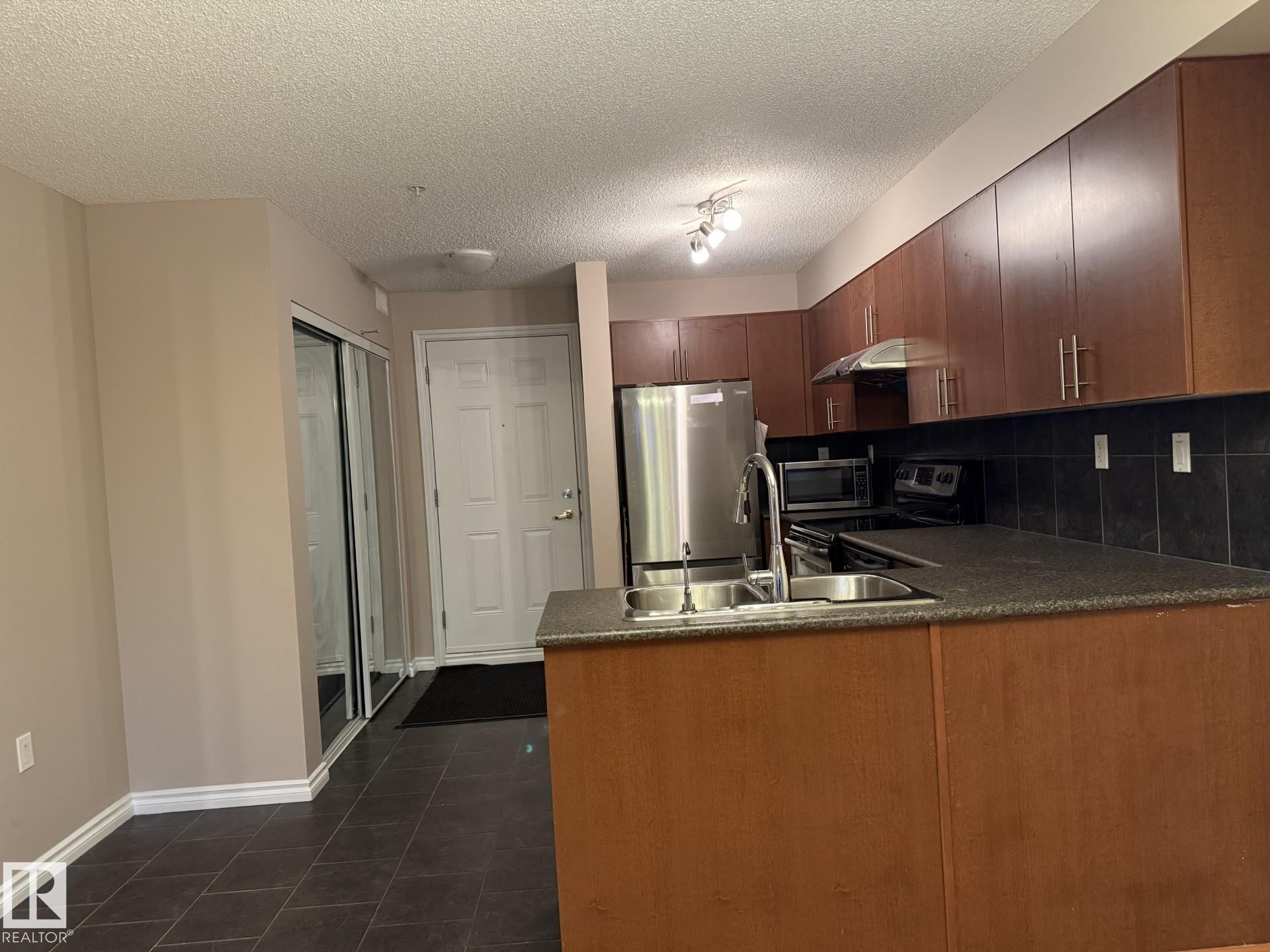 Kitchen with decorative backsplash, stainless steel appliances, a peninsula, dark tile patterned floors, and a textured ceiling - 303 10118 106 Avenue, Edmonton, AB - Indoor Photo Showing Kitchen With Double Sink