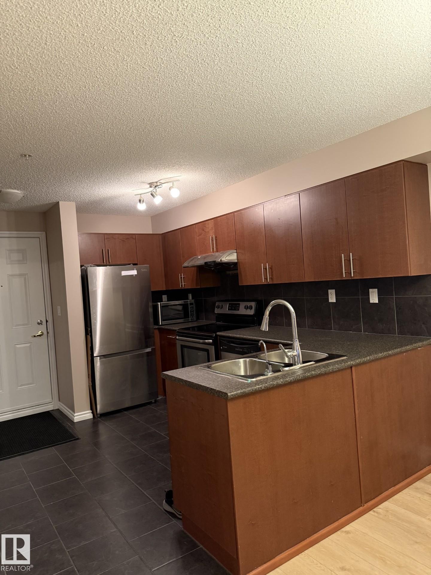 Kitchen featuring stainless steel appliances, a peninsula, tasteful backsplash, brown cabinets, and a textured ceiling - 303 10118 106 Avenue, Edmonton, AB - Indoor Photo Showing Kitchen With Double Sink