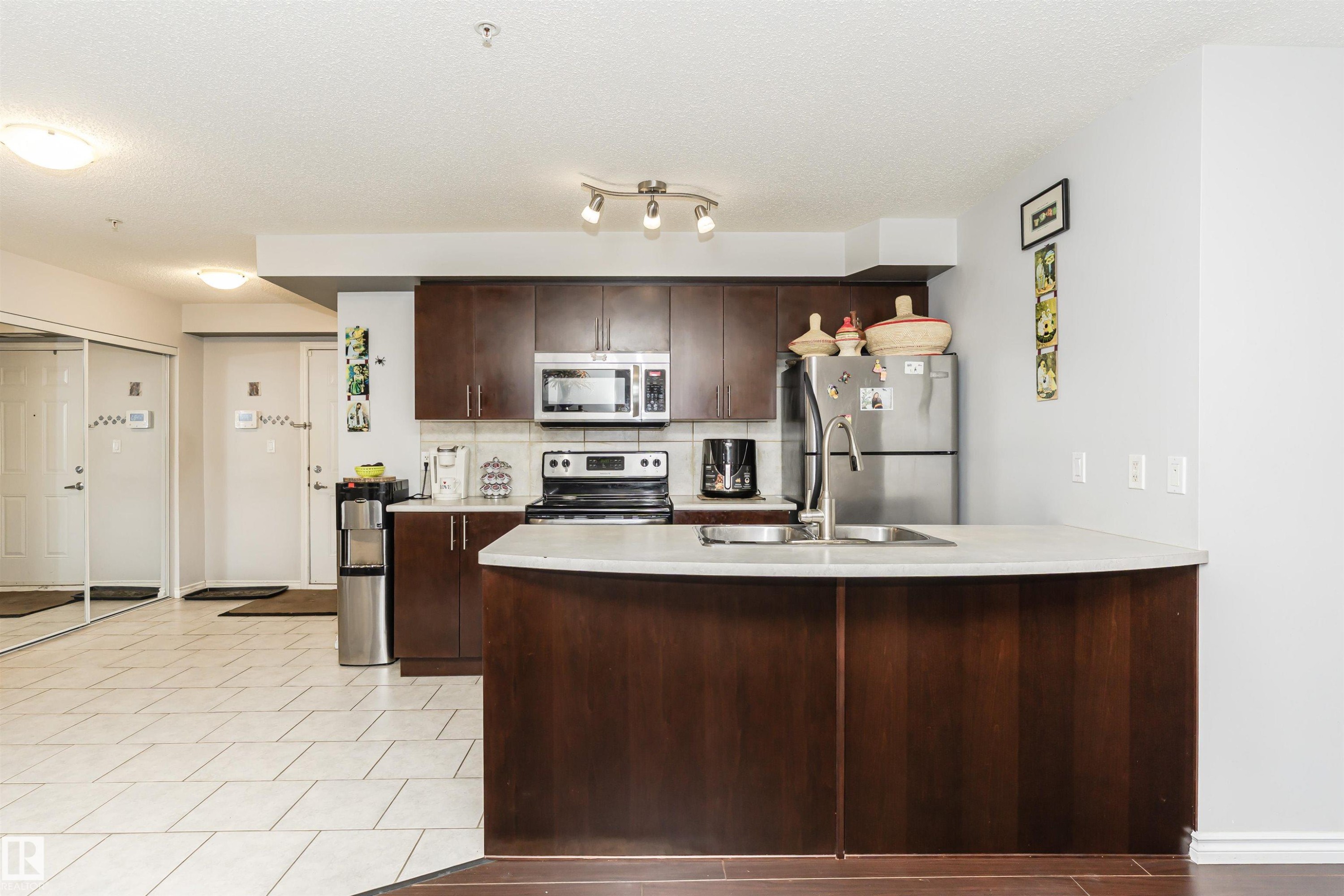 Kitchen featuring dark brown cabinetry, a peninsula, light countertops, appliances with stainless steel finishes, and a textured ceiling - 107 10118 106 Avenue, Edmonton, AB - Indoor Photo Showing Kitchen With Double Sink