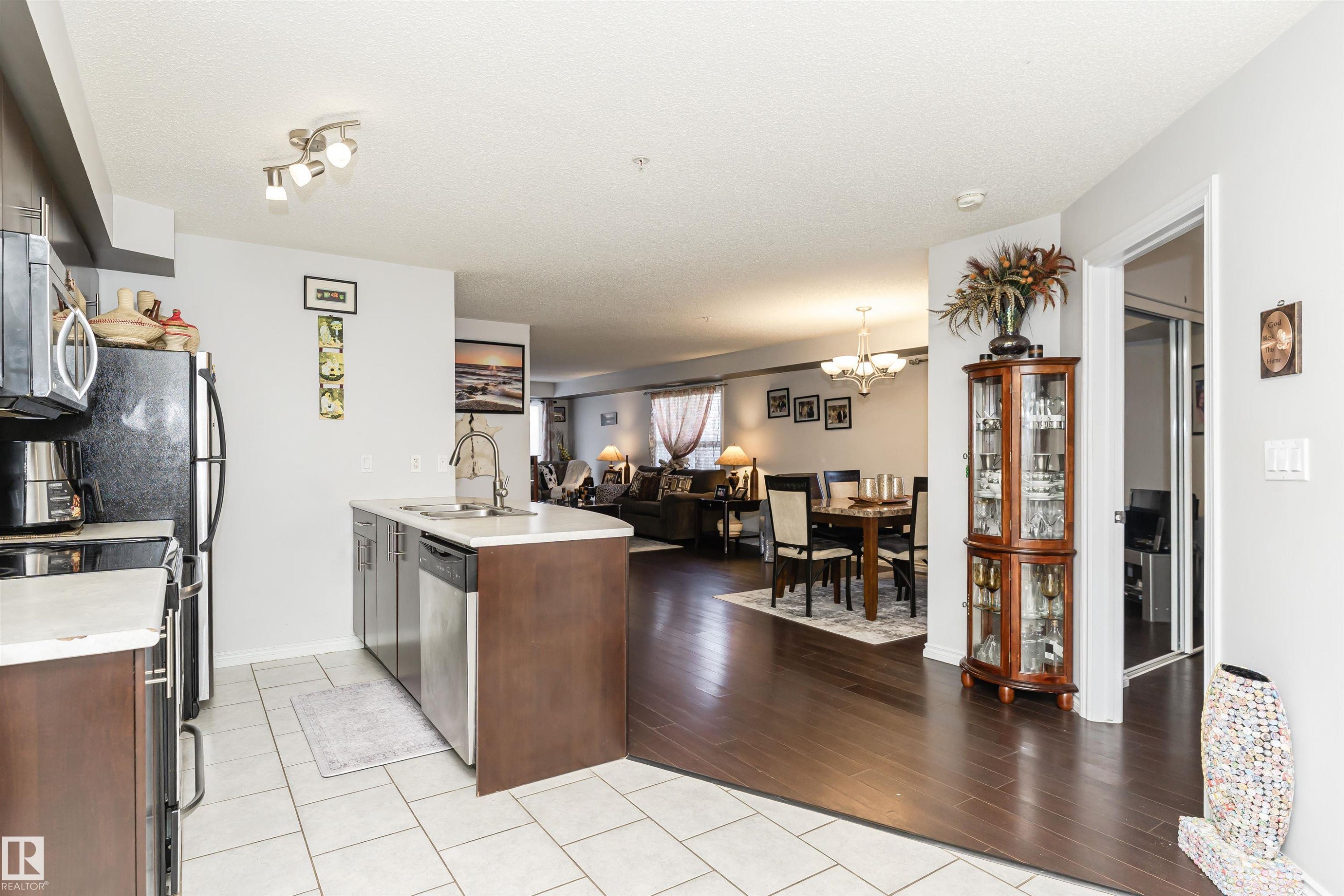 Kitchen featuring a peninsula, appliances with stainless steel finishes, open floor plan, light tile patterned floors, and a textured ceiling - 107 10118 106 Avenue, Edmonton, AB - Indoor Photo Showing Kitchen With Double Sink