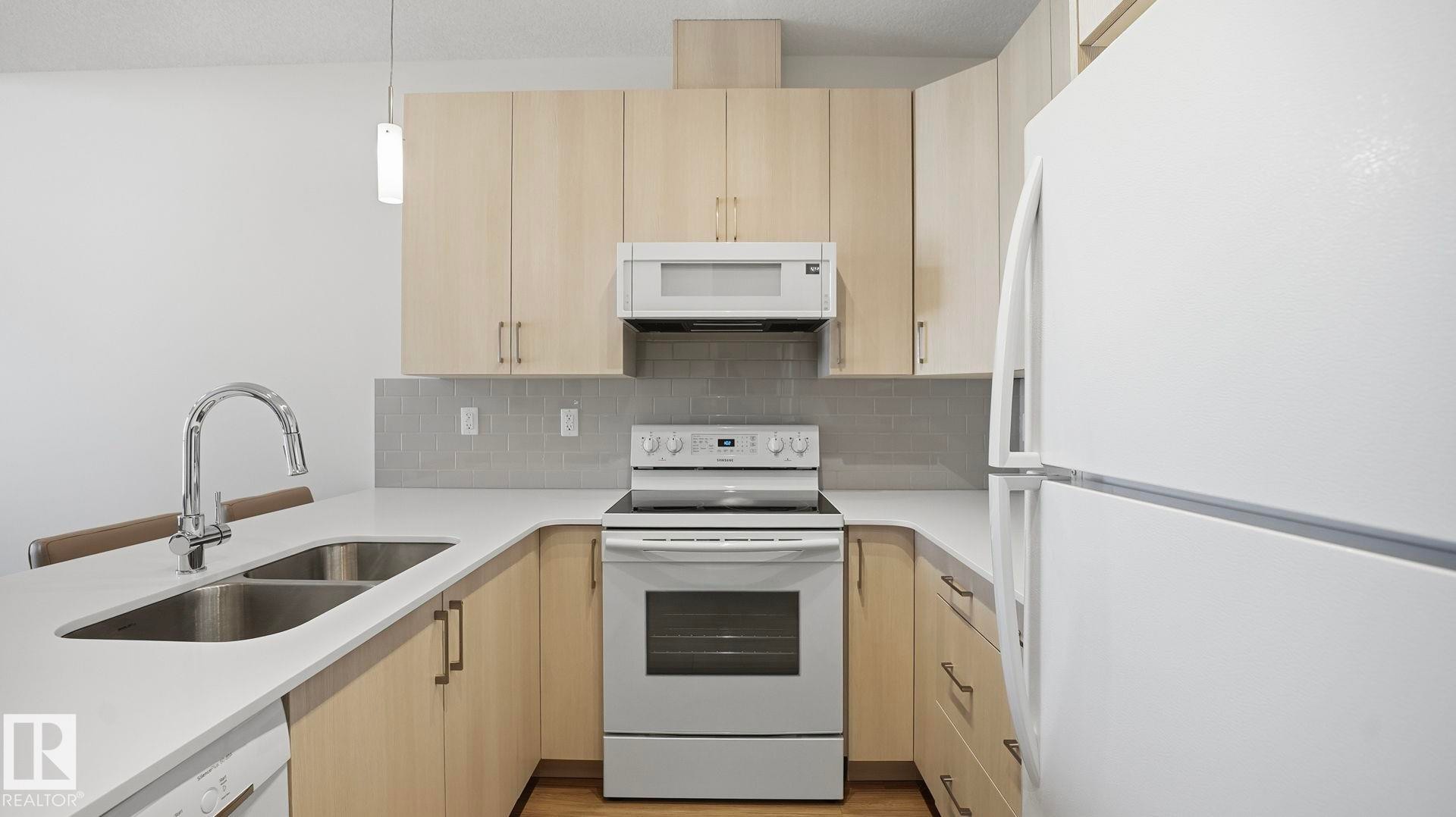 Kitchen with light brown cabinets, white appliances, decorative backsplash, hanging light fixtures, and light stone counters - 401 17 Columbia Avenue W, Devon, AB - Indoor Photo Showing Kitchen With Double Sink