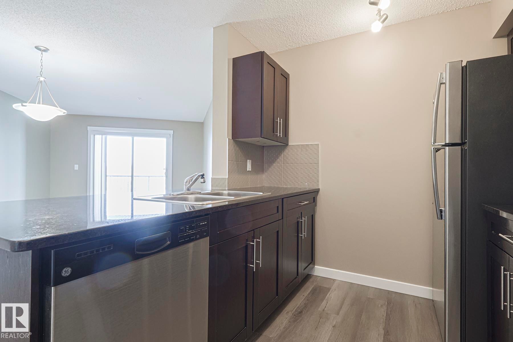Kitchen featuring stainless steel appliances, dark wood-style flooring, pendant lighting, a peninsula, and backsplash - 313 1510 Watt Drive, Edmonton, AB - Indoor Photo Showing Kitchen With Double Sink