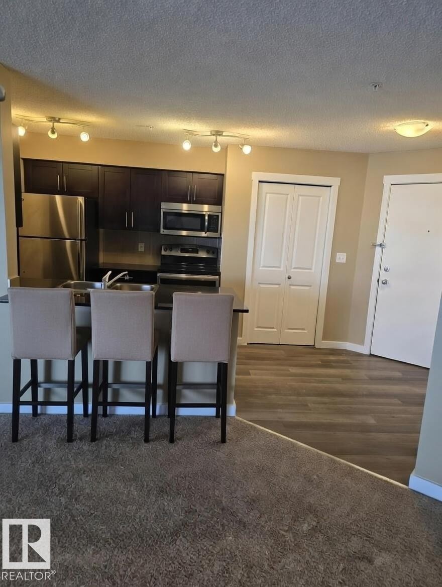 Kitchen featuring stainless steel appliances, rail lighting, a breakfast bar area, a textured ceiling, and dark countertops - 313 1510 Watt Drive, Edmonton, AB - Indoor Photo Showing Kitchen