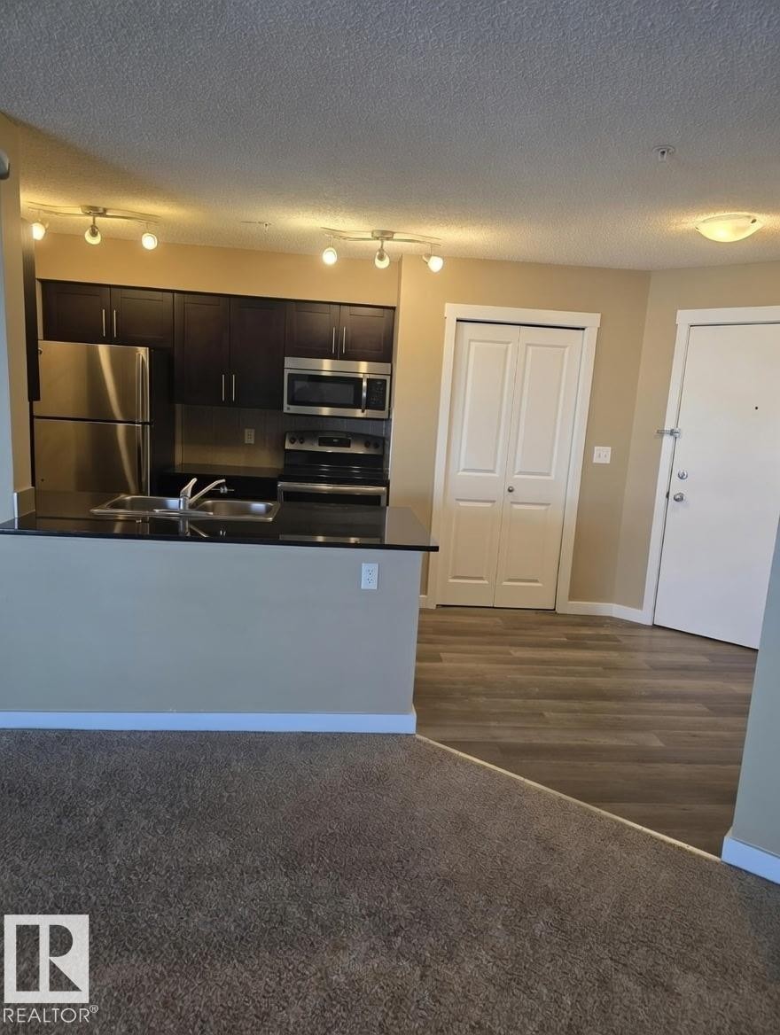 Kitchen with dark countertops, appliances with stainless steel finishes, track lighting, open floor plan, and a textured ceiling - 313 1510 Watt Drive, Edmonton, AB - Indoor Photo Showing Kitchen With Double Sink