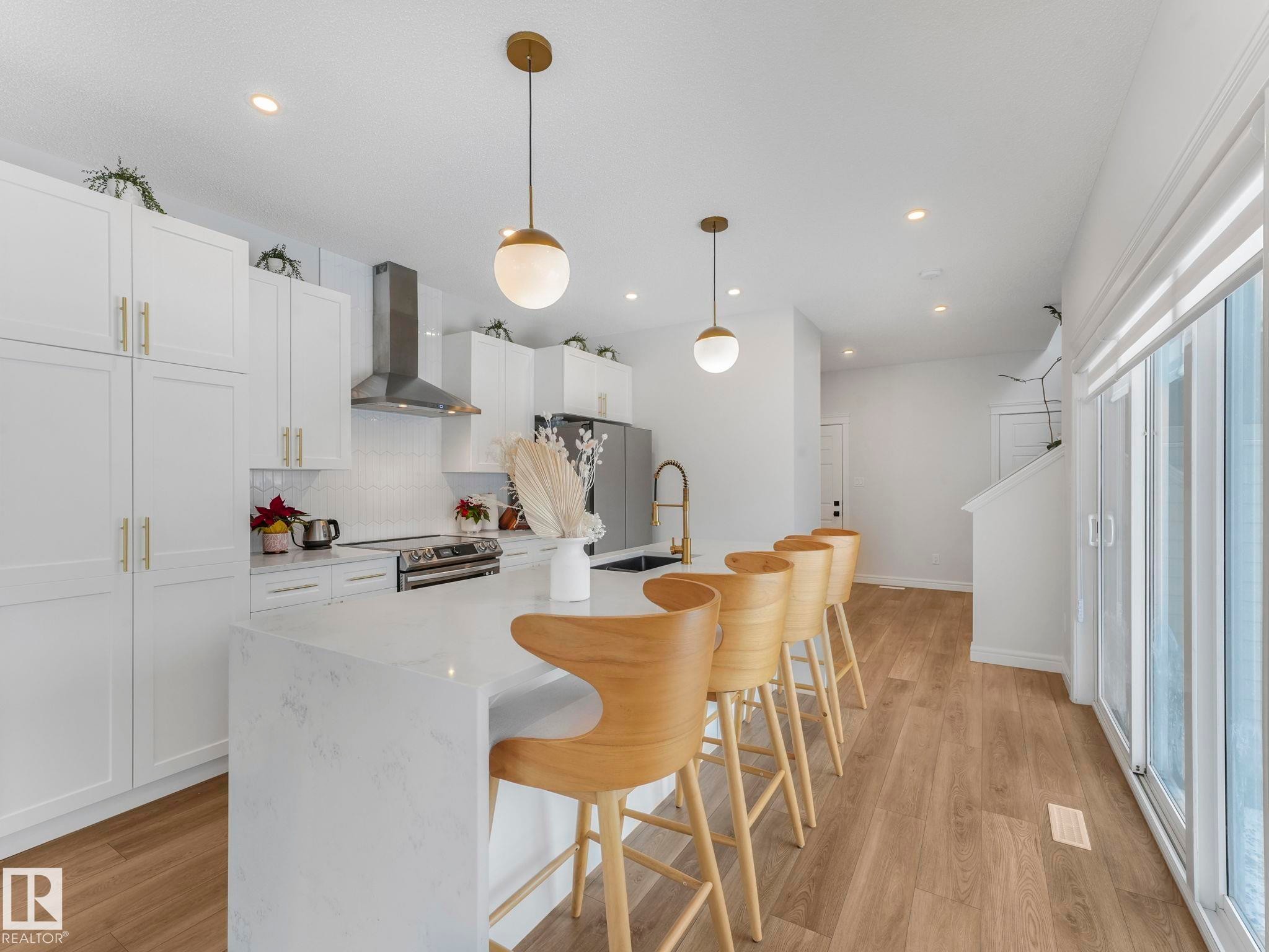 Kitchen featuring a breakfast bar area, decorative light fixtures, wall chimney range hood, light stone countertops, and backsplash - 4447 Kinsella Green, Edmonton, AB - Indoor Photo Showing Kitchen With Upgraded Kitchen