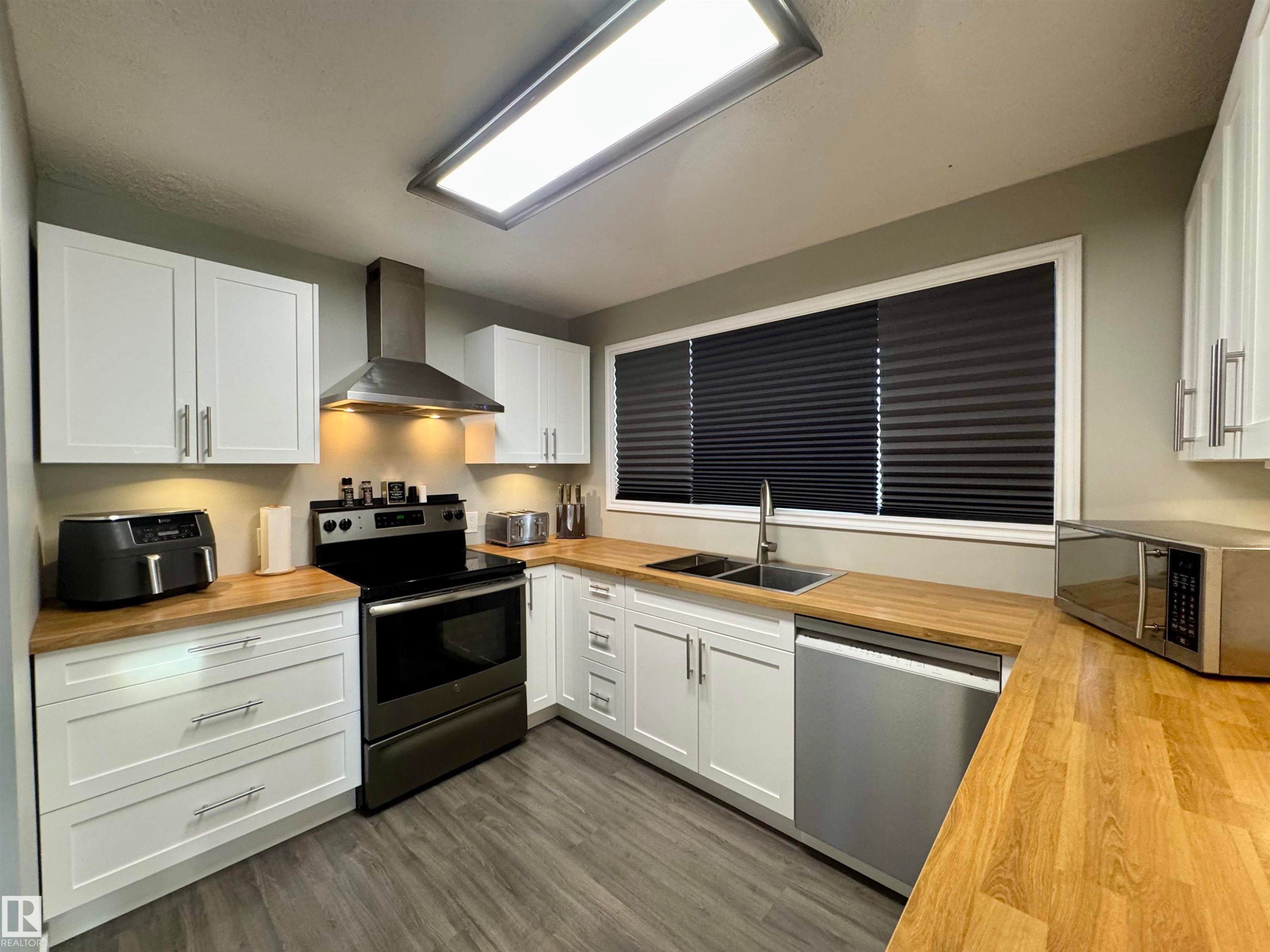 Kitchen featuring butcher block counters, appliances with stainless steel finishes, white cabinetry, and wall chimney range hood - 5140 52 Avenue, Ryley, AB - Indoor Photo Showing Kitchen With Double Sink