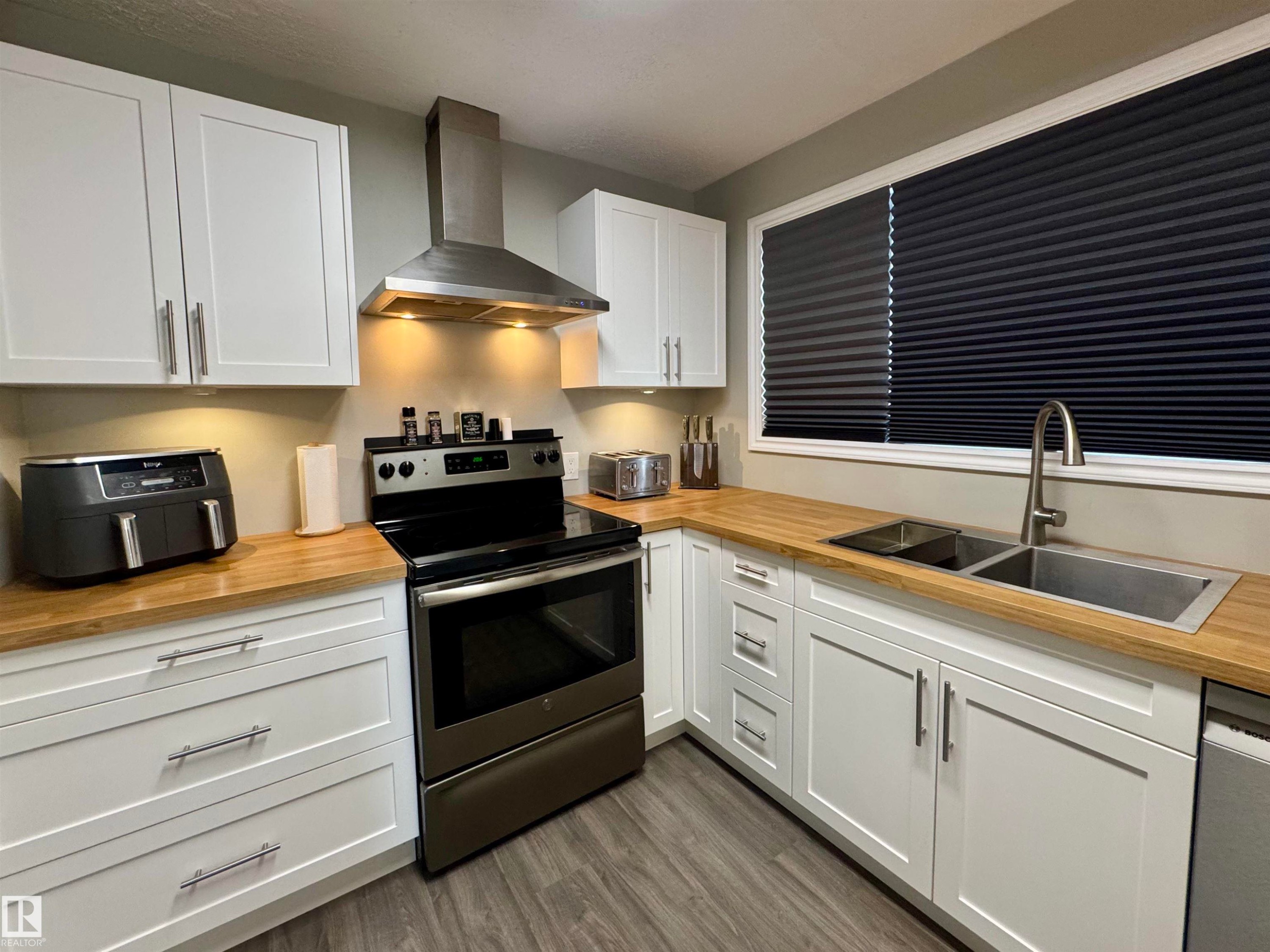Kitchen with appliances with stainless steel finishes, white cabinets, wall chimney exhaust hood, dark wood finished floors, and wood counters - 5140 52 Avenue, Ryley, AB - Indoor Photo Showing Kitchen With Double Sink