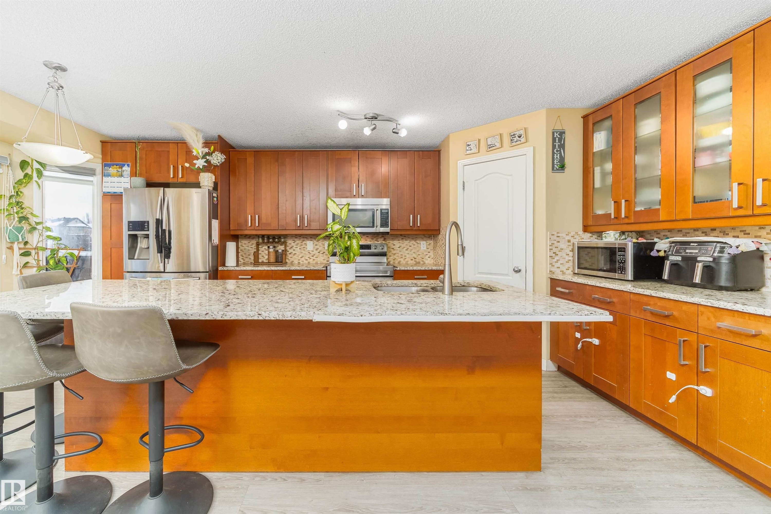 Kitchen featuring glass insert cabinets, an island with sink, backsplash, appliances with stainless steel finishes, and brown cabinets - 8518 6 Av, Edmonton, AB - Indoor Photo Showing Kitchen
