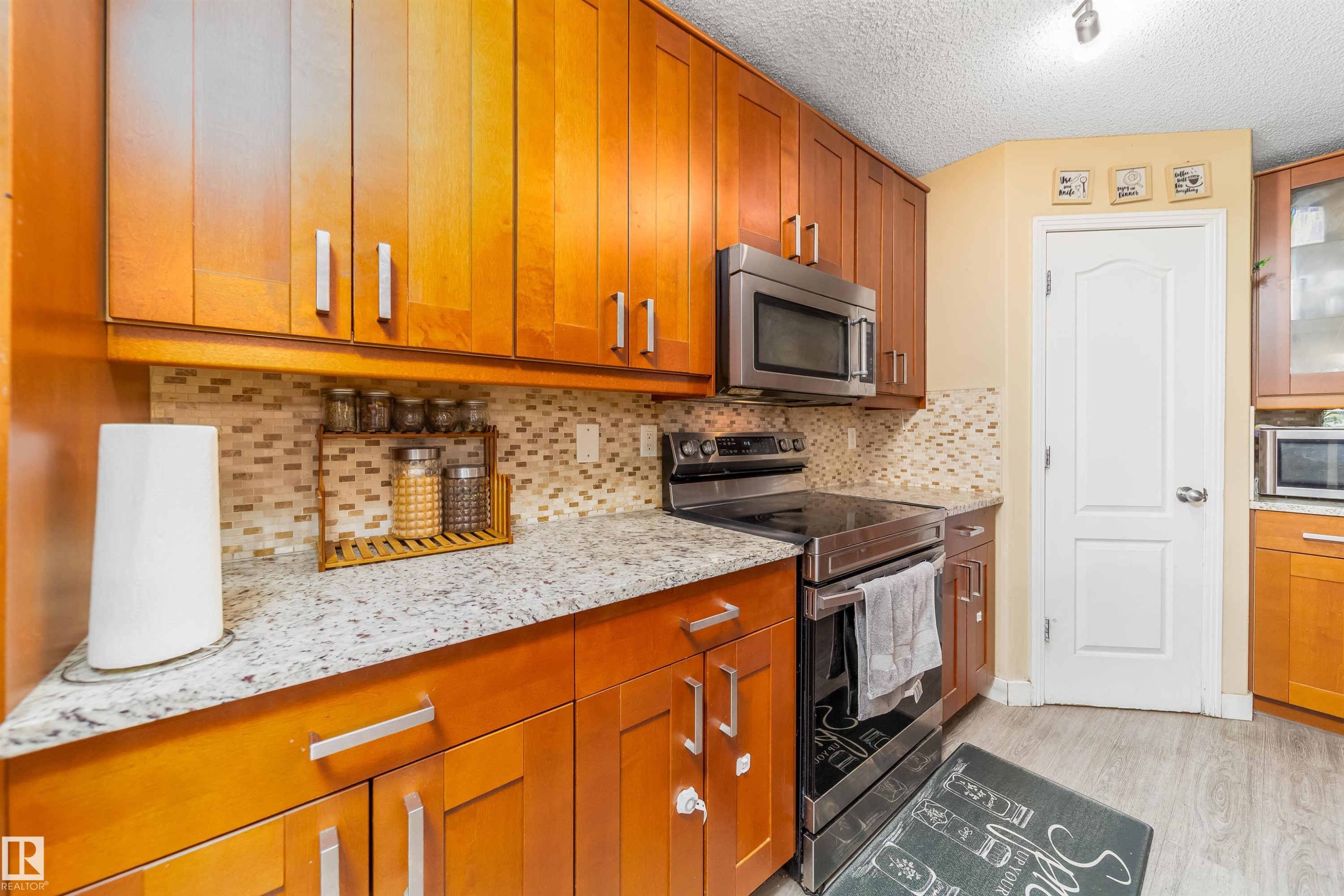 Kitchen featuring stainless steel appliances, brown cabinets, light stone countertops, light wood-style flooring, and a textured ceiling - 8518 6 Av, Edmonton, AB - Indoor Photo Showing Kitchen