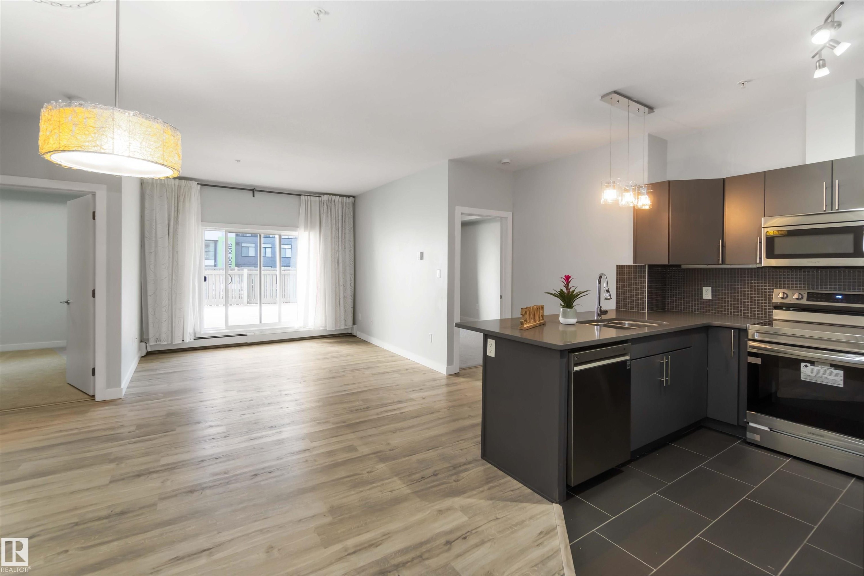 Kitchen featuring stainless steel appliances, hanging light fixtures, dark countertops, tasteful backsplash, and open floor plan - 107 10611 117 Street, Edmonton, AB - Indoor Photo Showing Kitchen