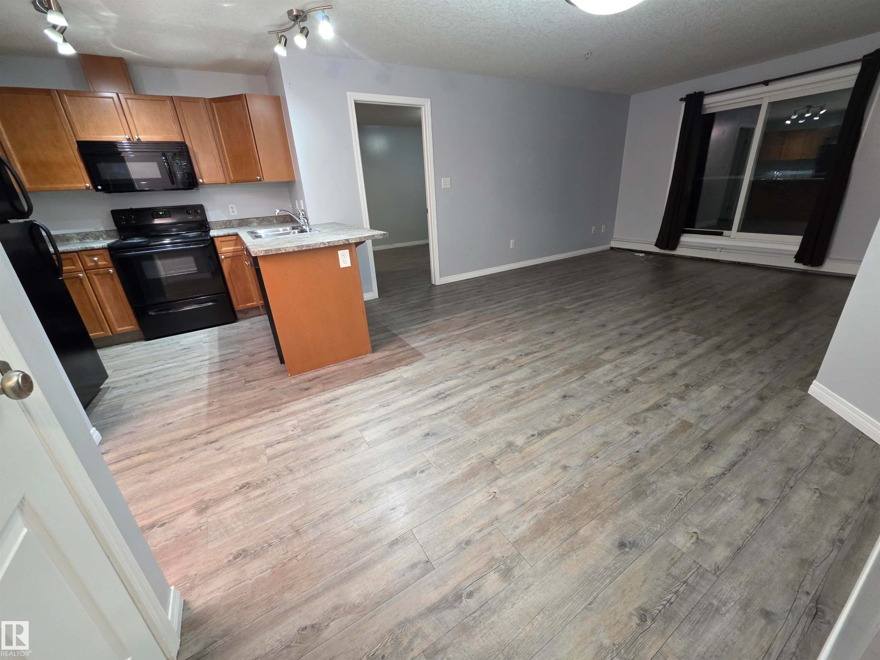 Kitchen with black appliances, open floor plan, light wood-type flooring, brown cabinetry, and a textured ceiling - 106 4903 47 Avenue, Stony Plain, AB - Indoor Photo Showing Kitchen With Double Sink