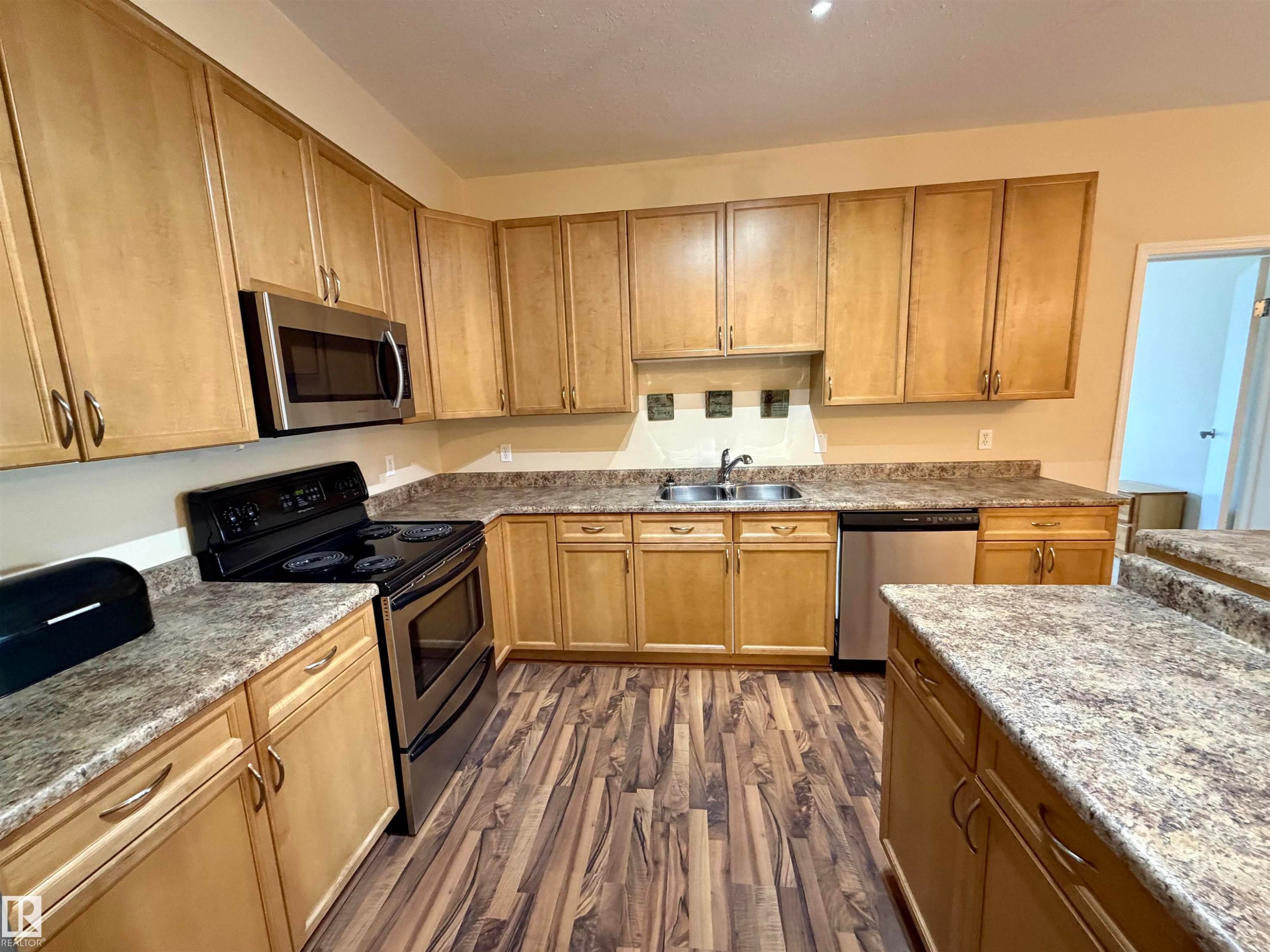 Kitchen featuring appliances with stainless steel finishes, dark wood finished floors, light stone countertops, and brown cabinetry - 321 4304 139 Avenue, Edmonton, AB - Indoor Photo Showing Kitchen With Double Sink