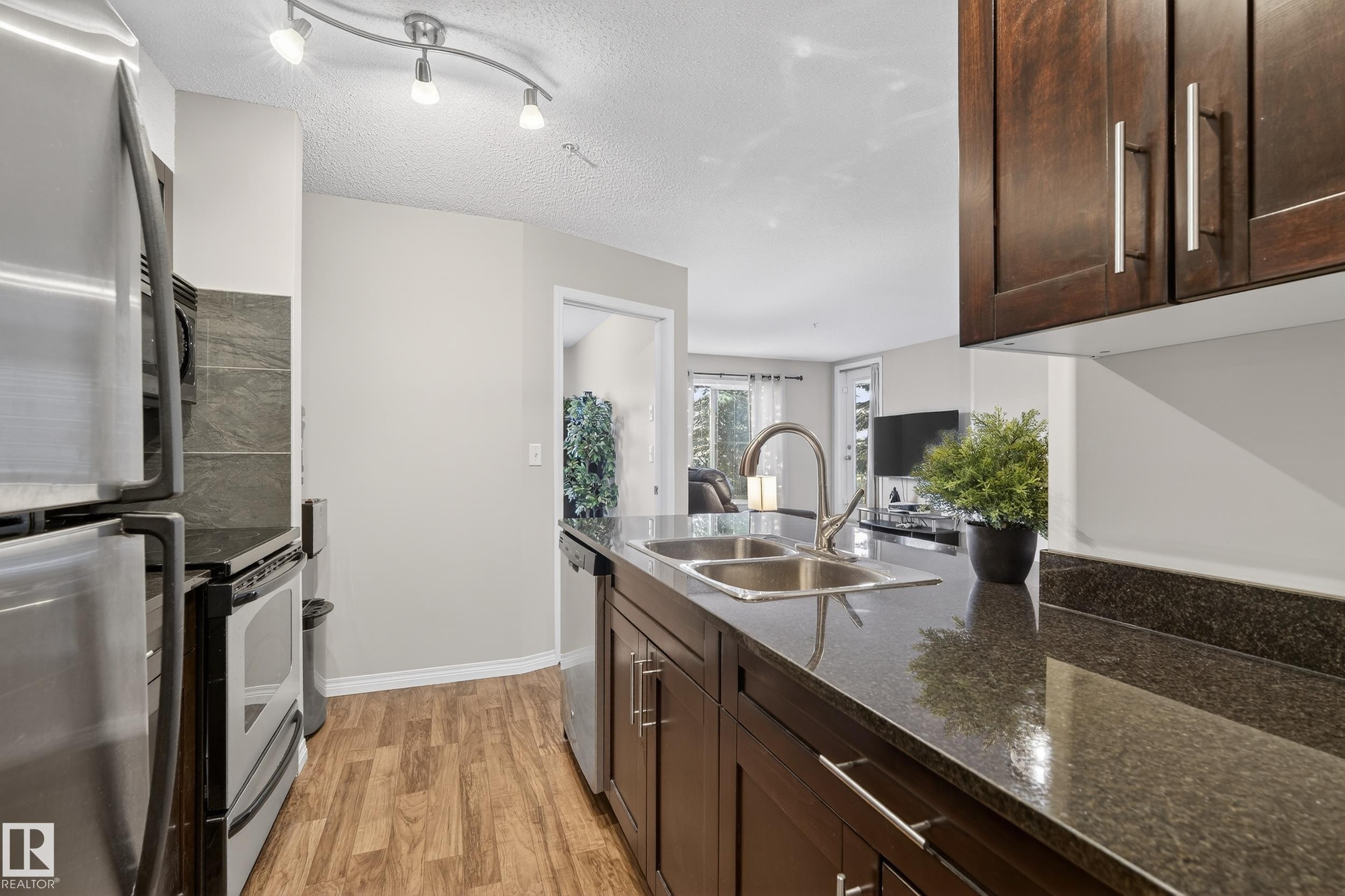 Kitchen featuring dark brown cabinets, stainless steel appliances, dark stone counters, light wood-type flooring, and a textured ceiling - 108 534 Watt Boulevard, Edmonton, AB - Indoor Photo Showing Kitchen With Double Sink