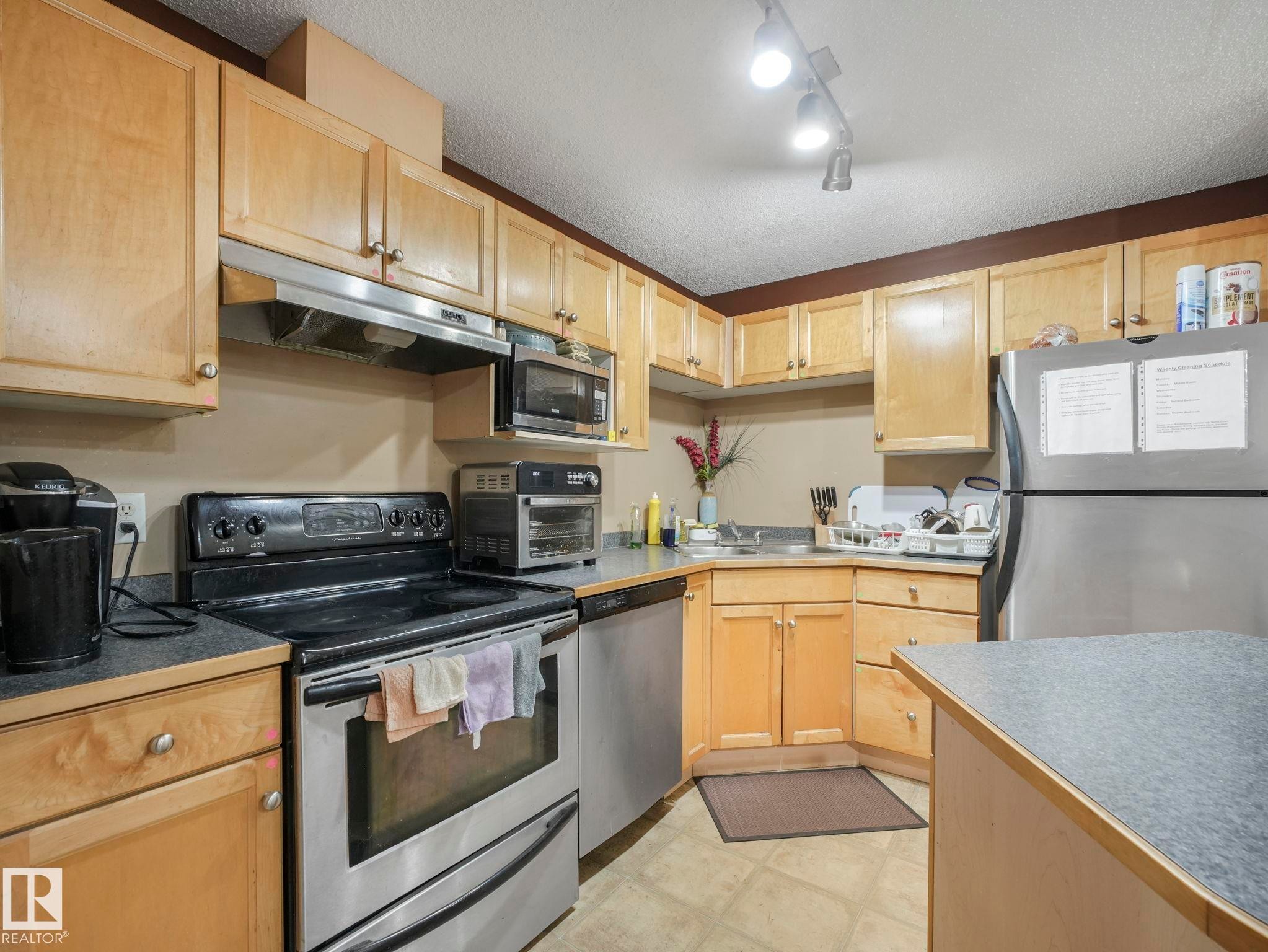 Kitchen featuring stainless steel appliances, a textured ceiling, under cabinet range hood, light brown cabinets, and light countertops - 210 245 Edwards Drive, Edmonton, AB - Indoor Photo Showing Kitchen