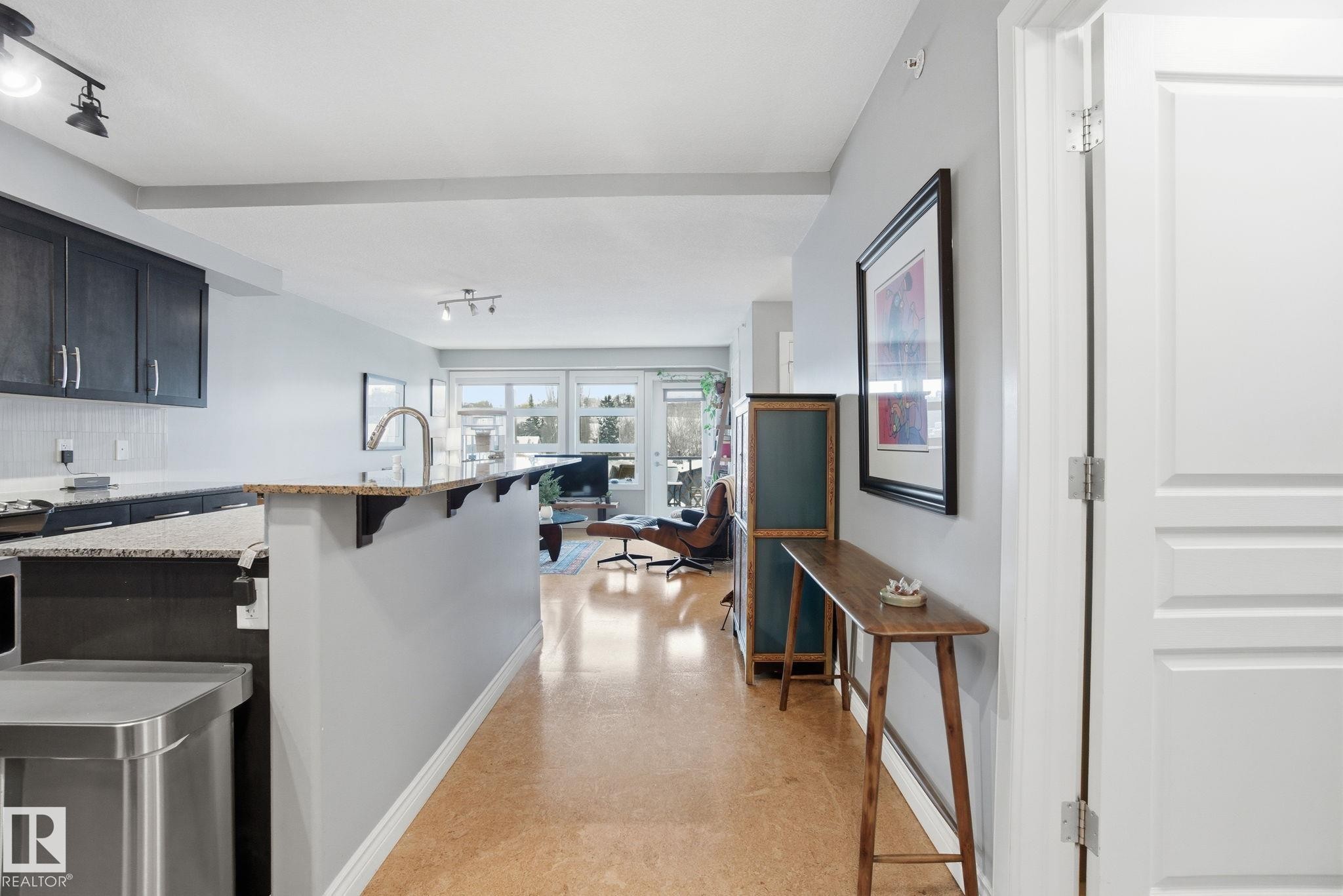 Kitchen featuring a breakfast bar, light stone counters, track lighting, a center island with sink, and dark cabinetry - 301 9603 98 Avenue, Edmonton, AB - Indoor