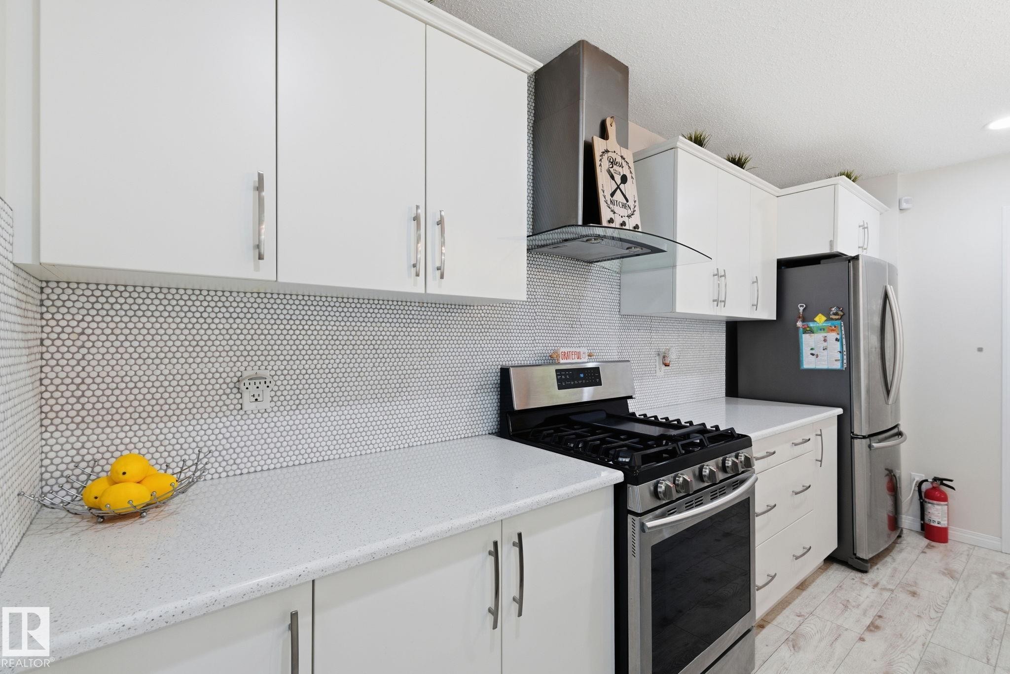 Kitchen featuring stainless steel appliances, wall chimney range hood, backsplash, white cabinetry, and light stone counters - 1511 Secord Rd, Edmonton, AB - Indoor Photo Showing Kitchen