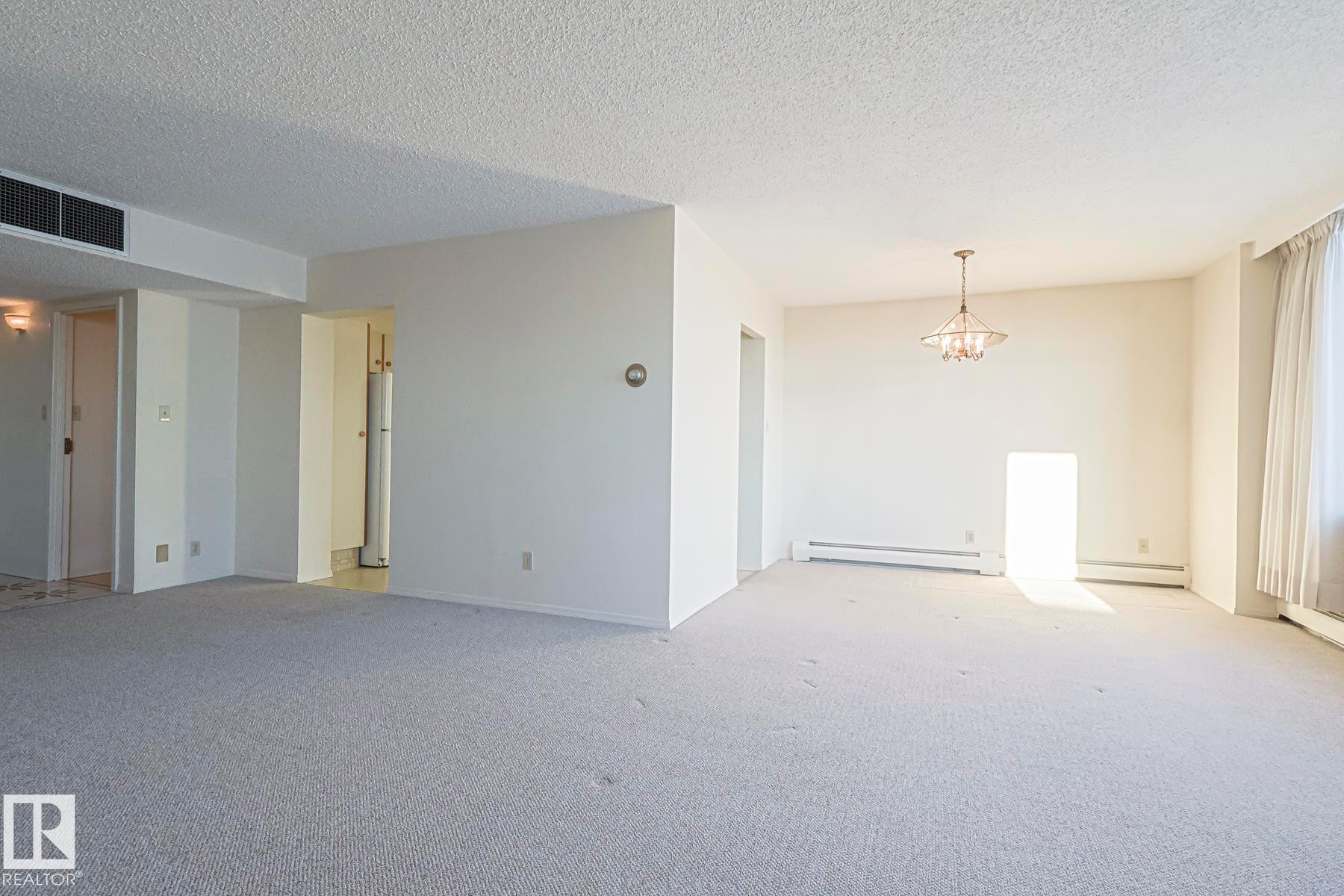 Spare room featuring a textured ceiling, a chandelier, a baseboard heating unit, and light carpet - 302 8340 Jasper Avenue Nw, Edmonton, AB - Indoor Photo Showing Other Room