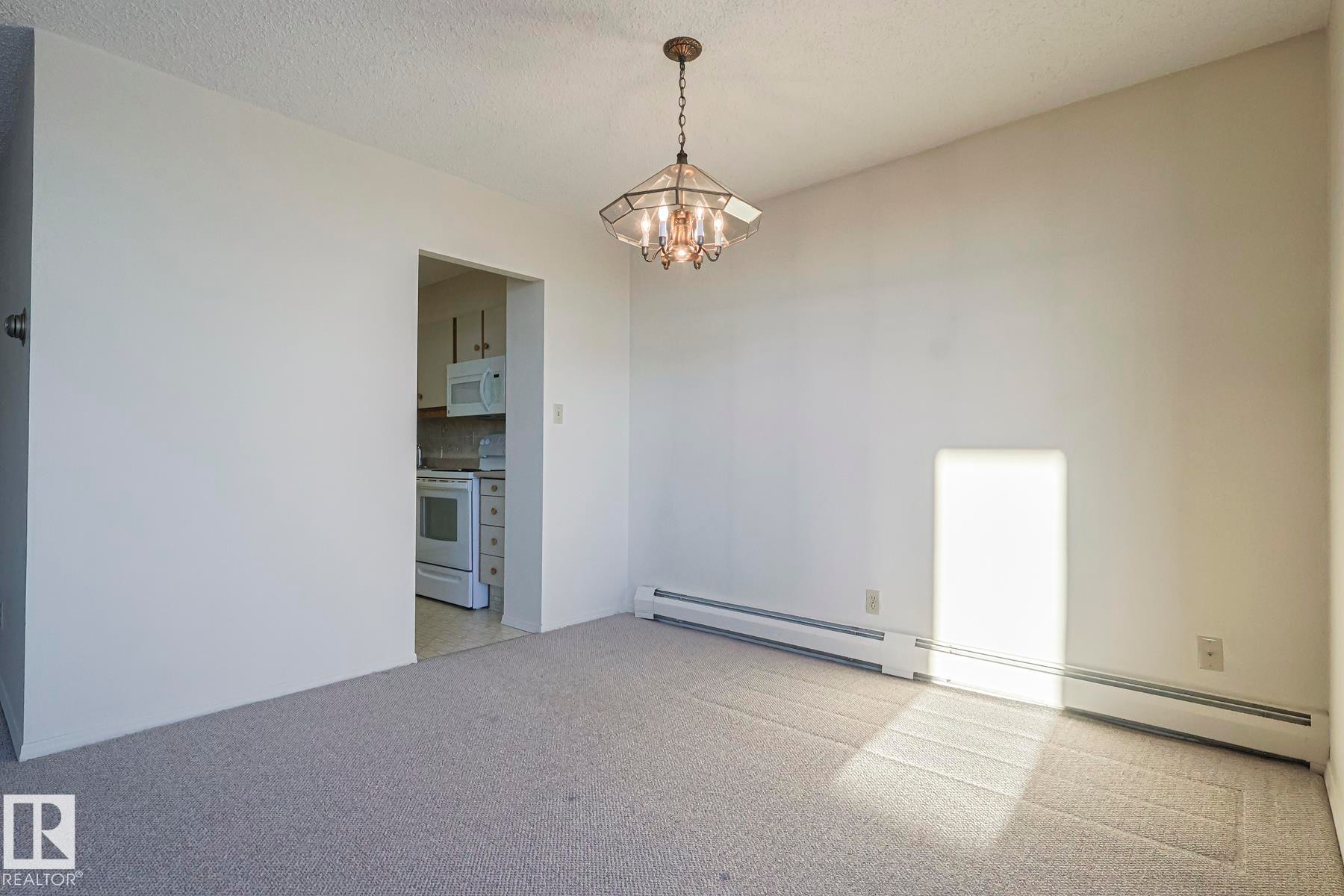 Empty room with baseboard heating, light colored carpet, a chandelier, and a textured ceiling - 302 8340 Jasper Avenue Nw, Edmonton, AB - Indoor Photo Showing Other Room