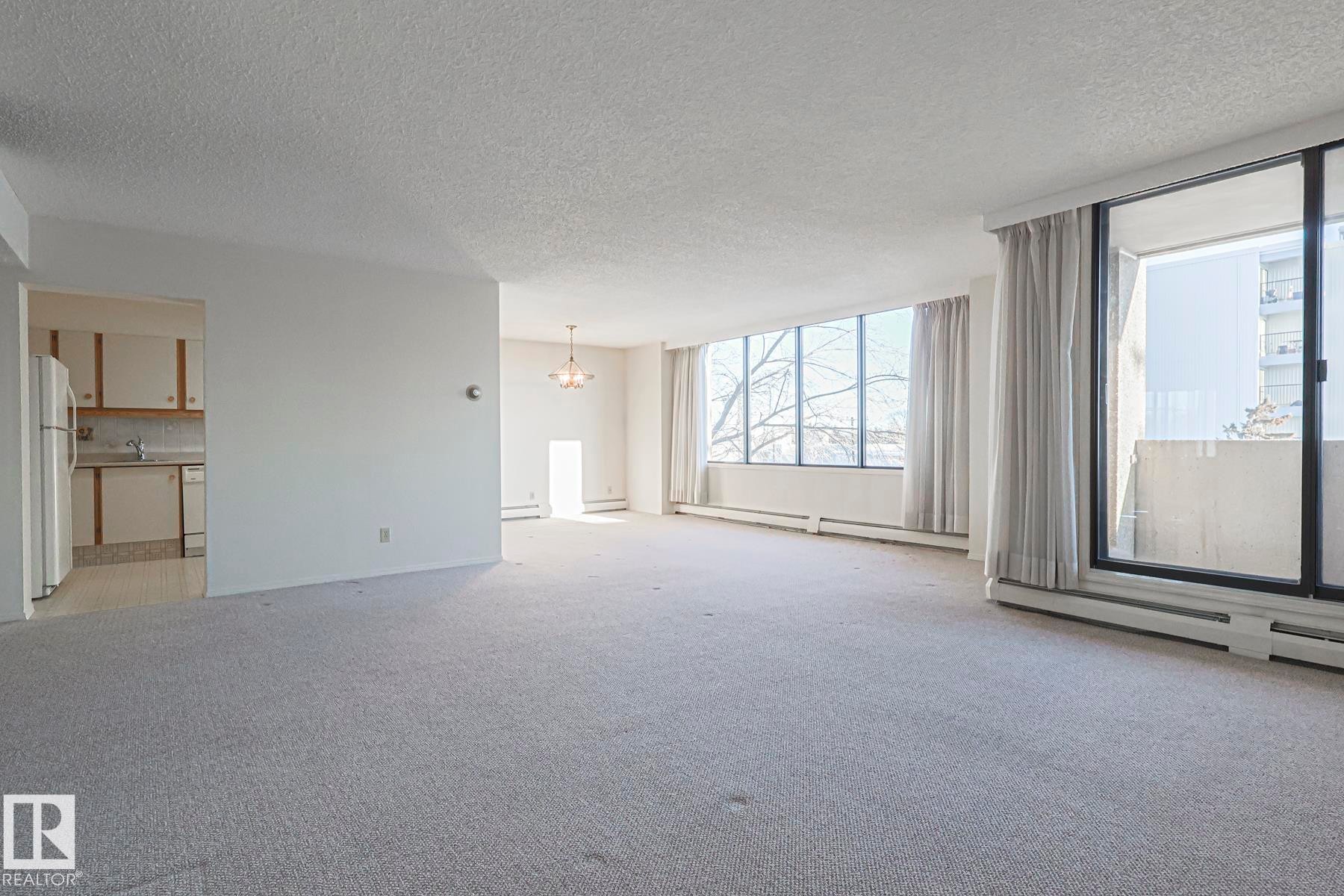 Unfurnished living room featuring a chandelier, a textured ceiling, light colored carpet, baseboard heating, and a baseboard heating unit - 302 8340 Jasper Avenue Nw, Edmonton, AB - Indoor Photo Showing Other Room