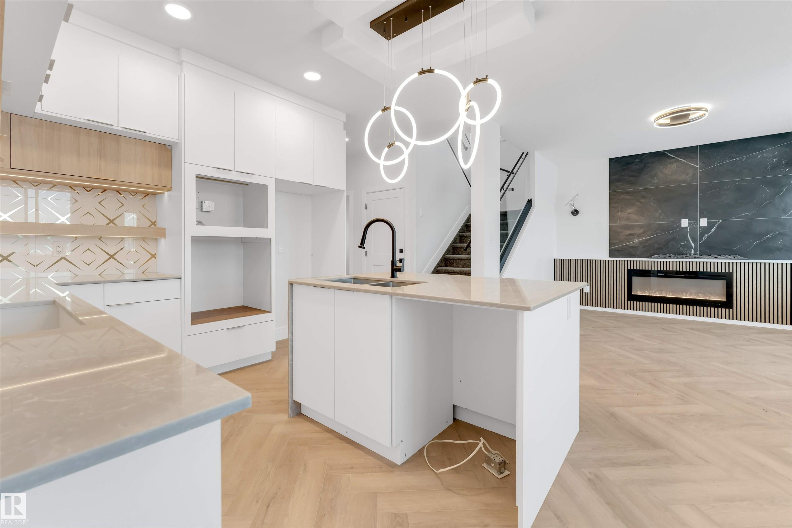 Kitchen featuring white cabinetry, an island with sink, light stone countertops, a glass covered fireplace, and modern cabinets - 150 Creekside Lane, Leduc, AB - Indoor Photo Showing Kitchen With Double Sink