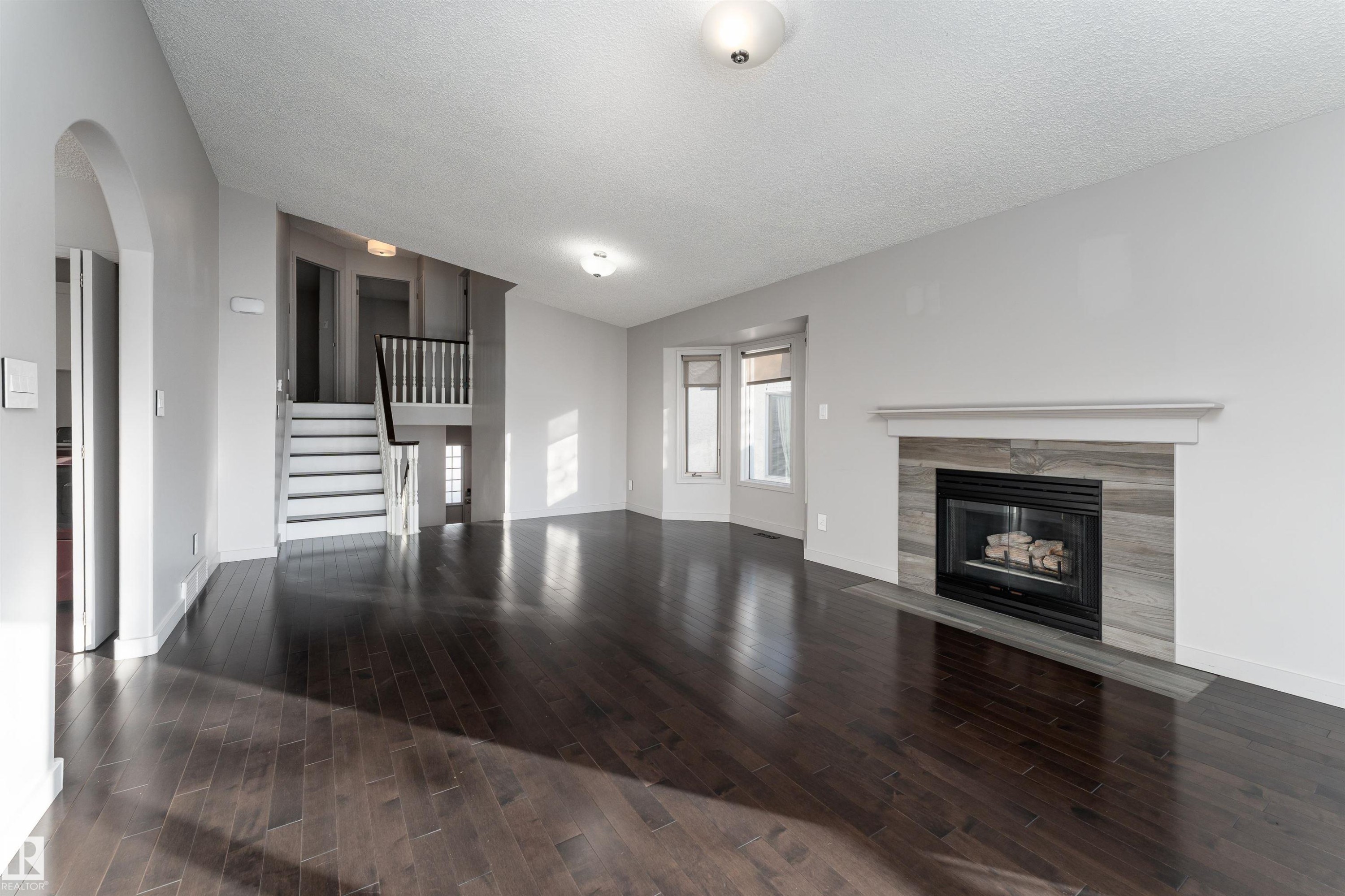 Unfurnished living room featuring lofted ceiling, a tiled fireplace, stairs, dark wood-type flooring, and arched walkways - 174 Oeming Road, Edmonton, AB - Indoor Photo Showing Living Room With Fireplace