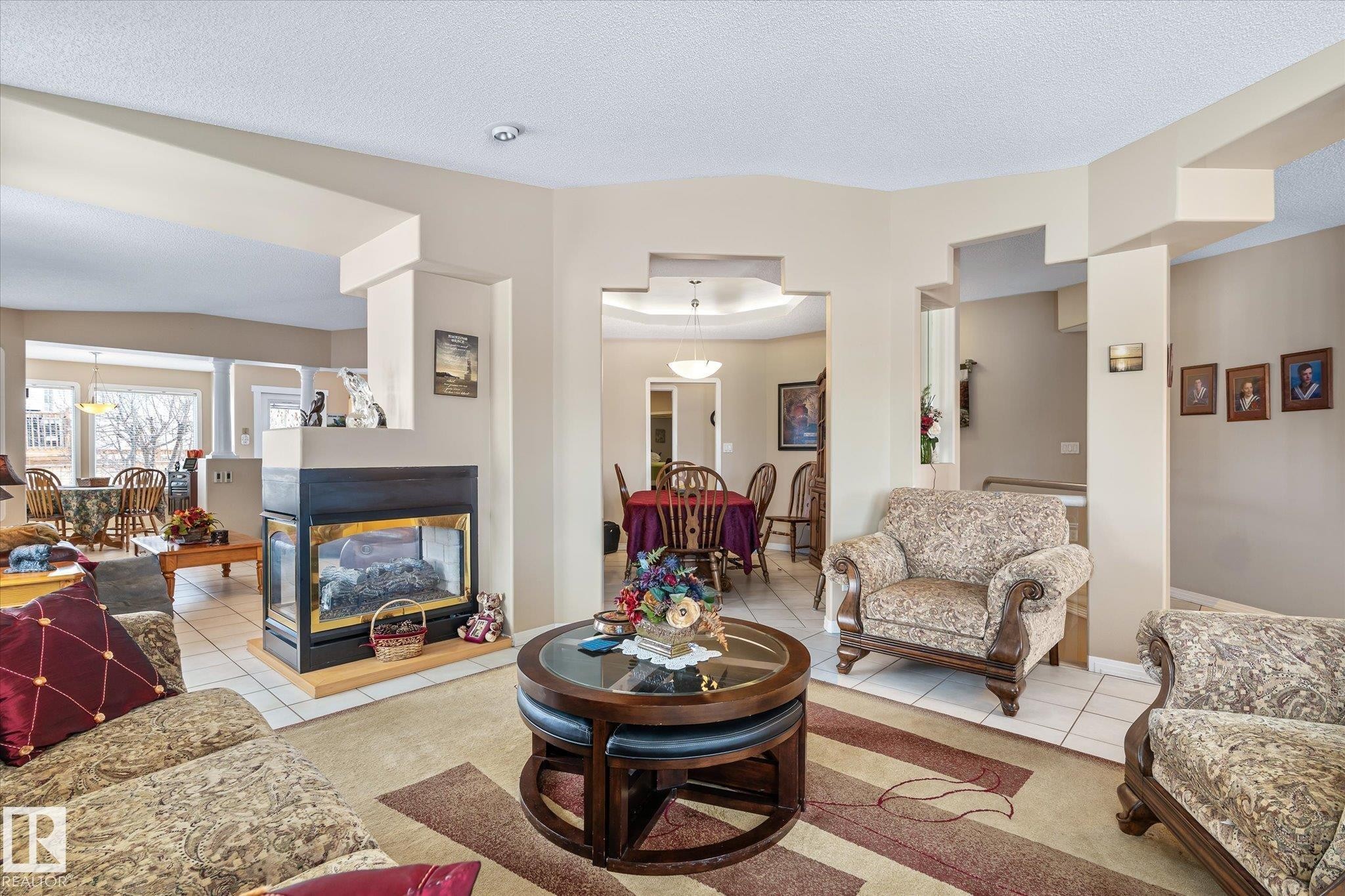 Living area with tile patterned flooring, a textured ceiling, a multi sided fireplace, carpet floors, and a raised ceiling - 7020 157 Avenue, Edmonton, AB - Indoor Photo Showing Living Room With Fireplace