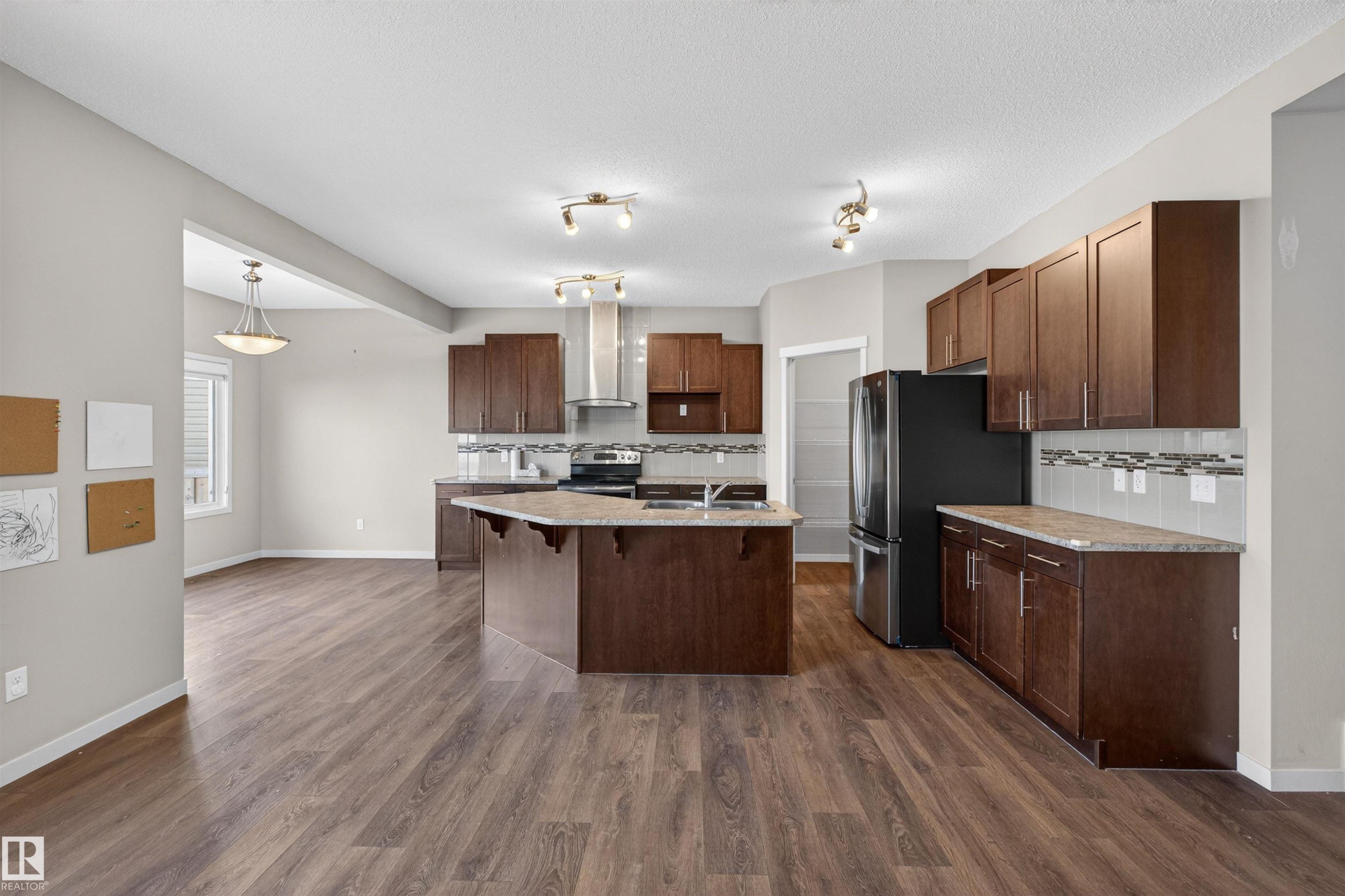 Kitchen with appliances with stainless steel finishes, an island with sink, a breakfast bar, wall chimney exhaust hood, and decorative light fixtures - 4723 Alwood Bend Bend Sw, Edmonton, AB - Indoor Photo Showing Kitchen