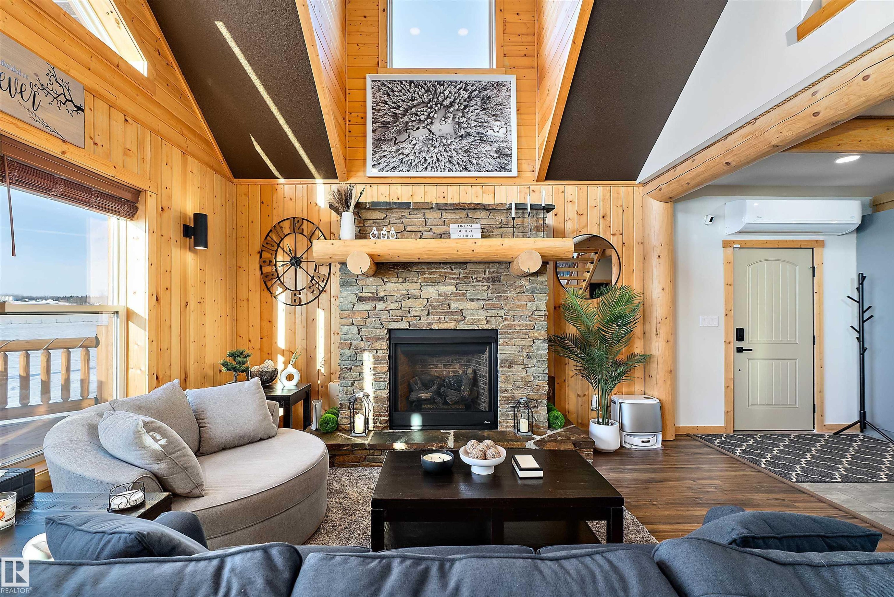 Living room featuring wood finished floors, a stone fireplace, wood walls, high vaulted ceiling, and a wall mounted air conditioner - 19003 Twp Rd 522, Rural Beaver County, AB - Indoor Photo Showing Living Room With Fireplace